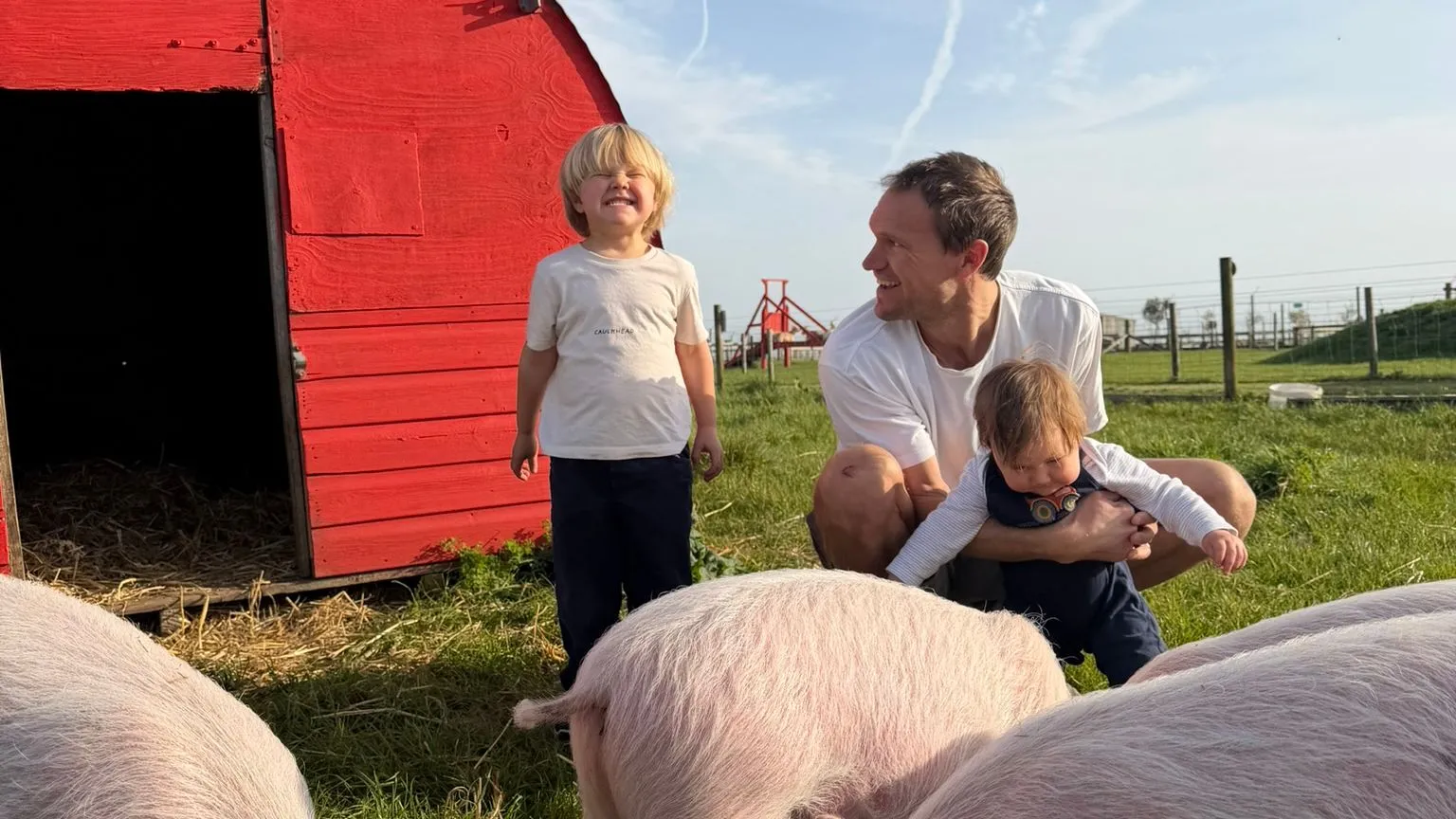 Adult and two children with pigs beside a bright red pig shelter in a grassy farm field.