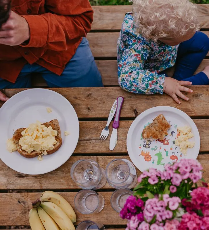 Overhead view of a family eating scrambled eggs on toast at a rustic outdoor wooden table.