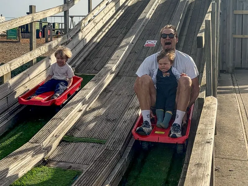 An adult and two young children ride red sledges down a wooden outdoor slide beneath a bright blue sky.
