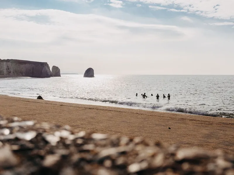 People swimming in the sea off a sandy beach with dramatic coastal rock stacks under a cloudy sky.