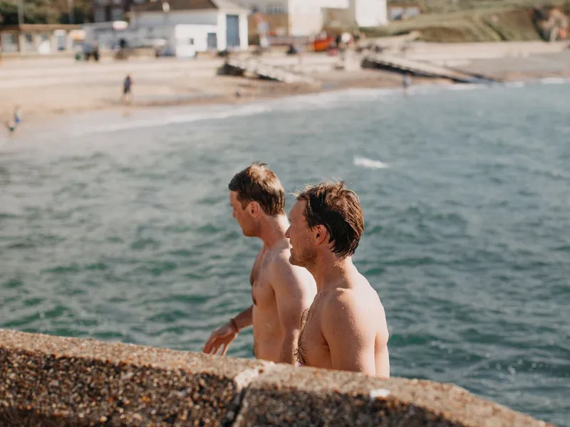 Three shirtless men standing by the sea beside a concrete harbour wall.