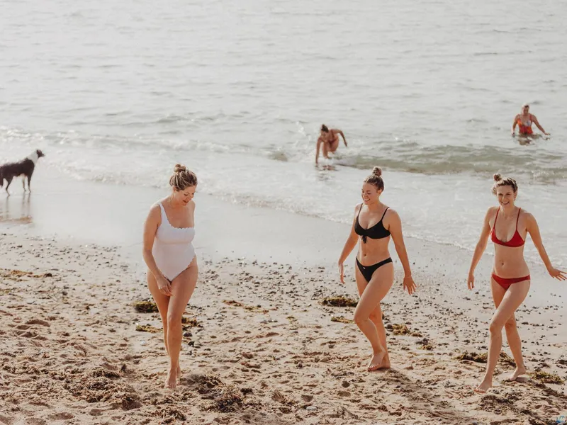 Three women in swimsuits walking up a sandy beach after swimming, with other bathers and a dog by the sea.