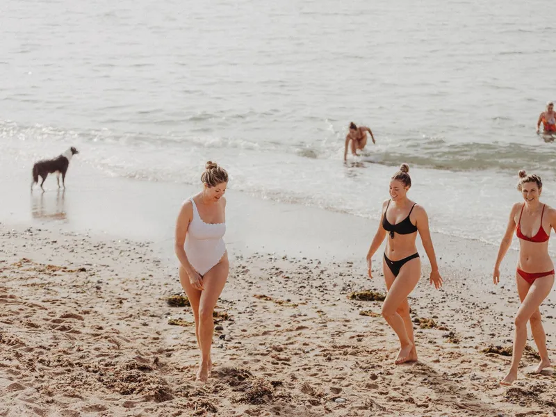 Three women in swimsuits walking up a sandy beach after swimming, with other bathers and a dog by the sea.
