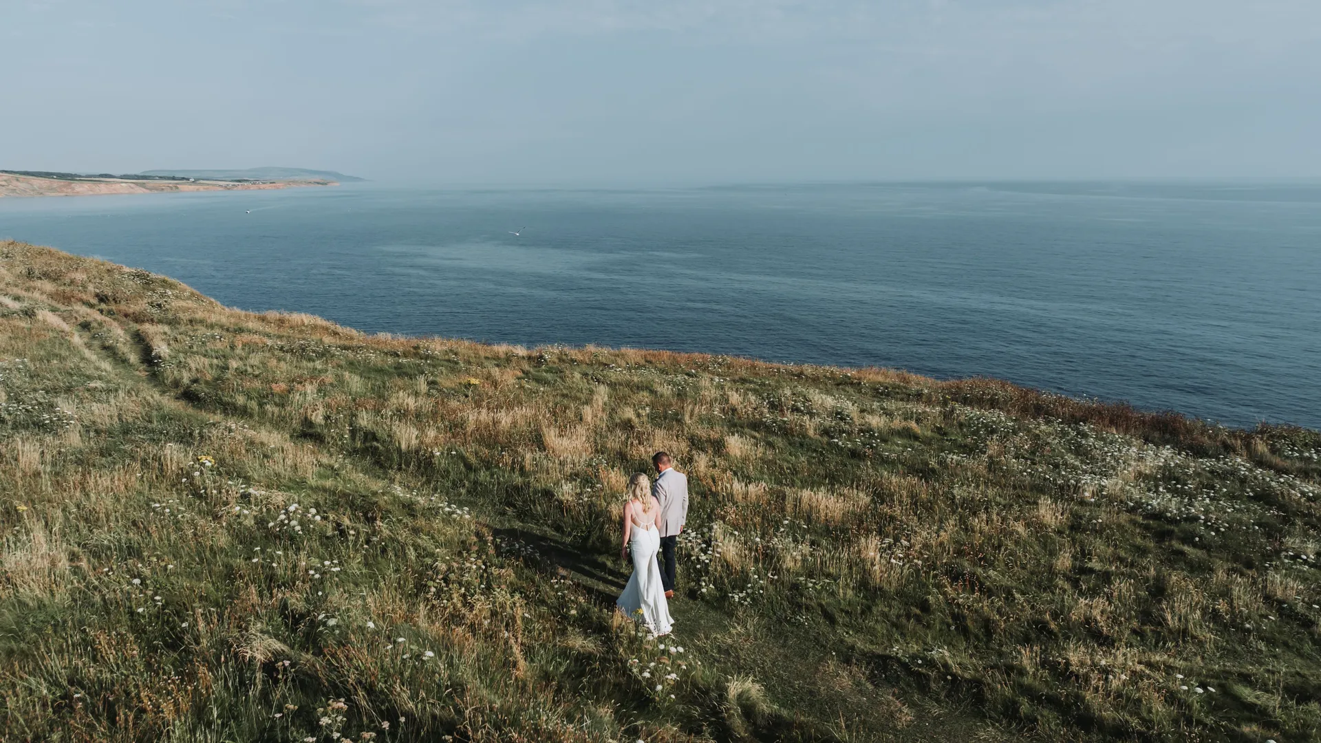 Newlywed couple walking through a wildflower meadow above a calm sea coast.