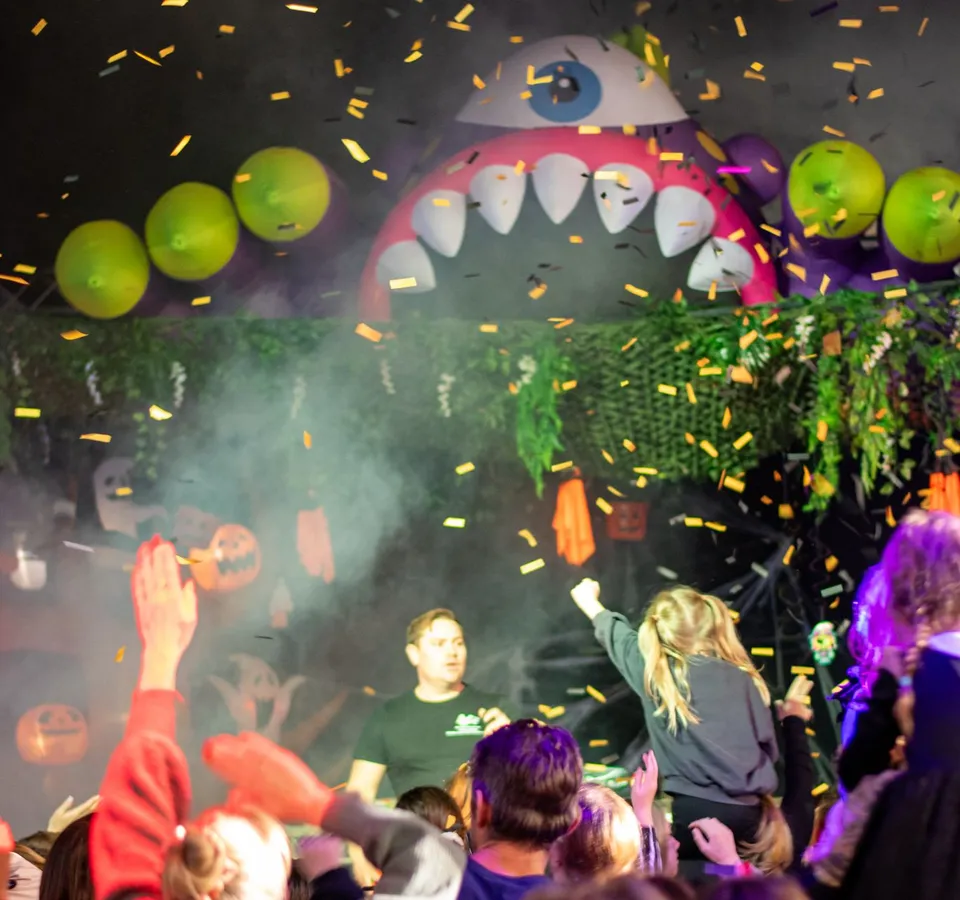 Crowd celebrating beneath falling confetti at a Halloween-themed event with a giant inflatable monster backdrop.