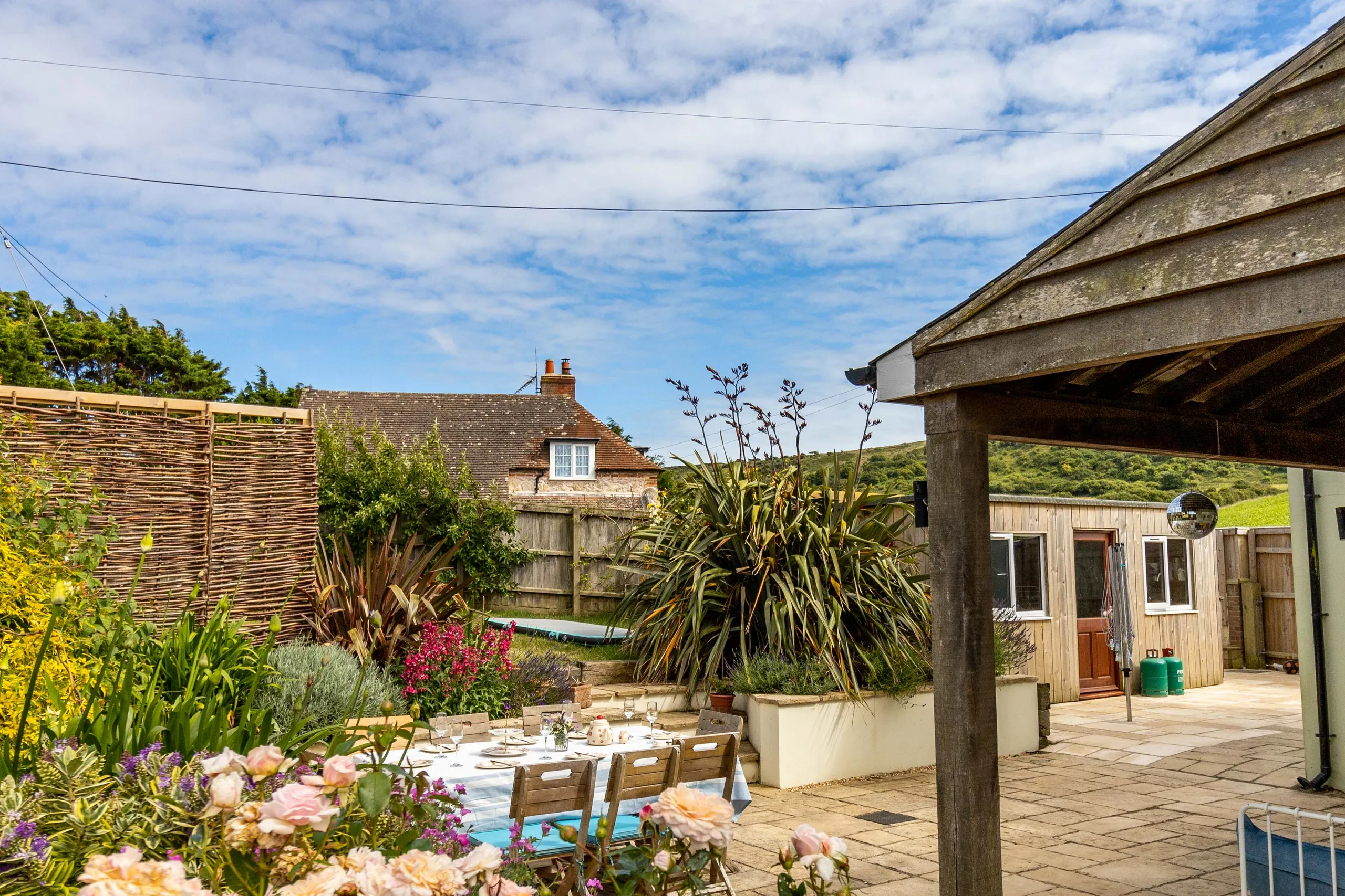 Outdoor patio dining area with table set for a meal, surrounded by colourful garden plants and a wooden outbuilding