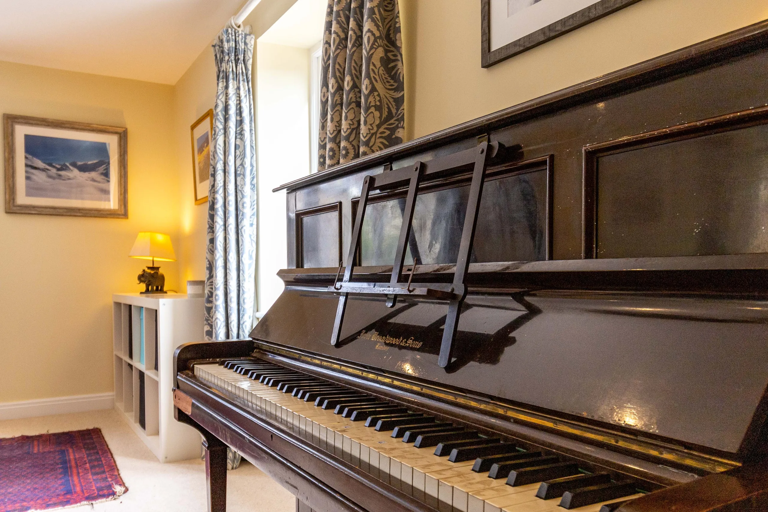 Upright piano in a cosy room with patterned curtains and a warm table lamp