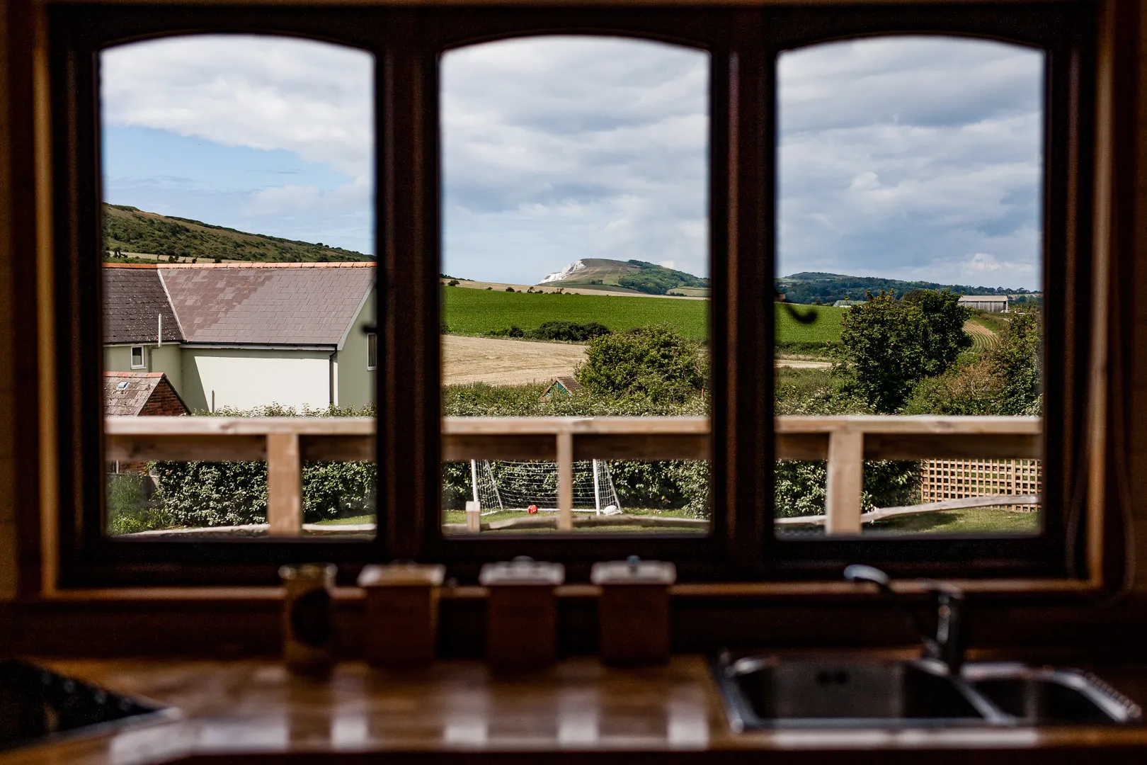 Countryside view through a three-panel window above a kitchen sink, with green fields and distant hills under a cloudy sky