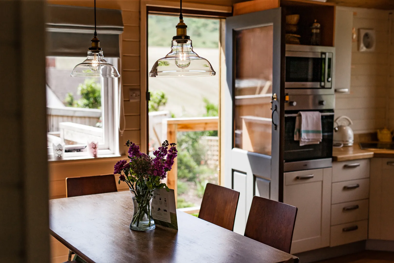 Cosy cabin kitchen-diner with wooden table, pendant lights and open door to a sunlit deck