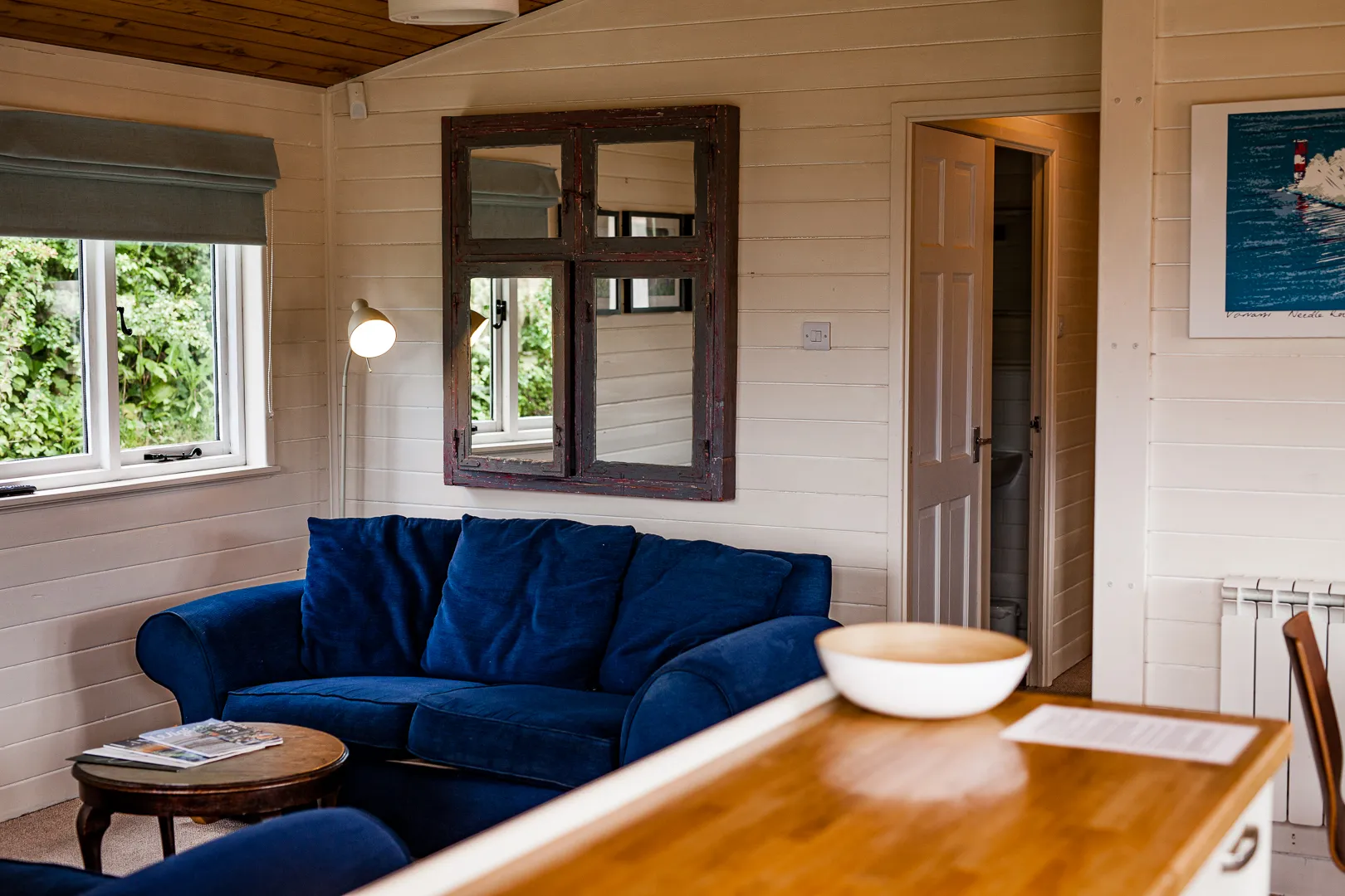Cosy cabin living area with a deep blue sofa, wooden ceiling, window with blind and a wall mirror