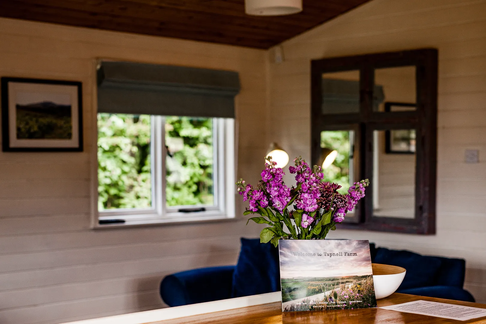 Cosy lodge living area with purple flowers on a wooden table, a blue sofa, and a window looking onto greenery