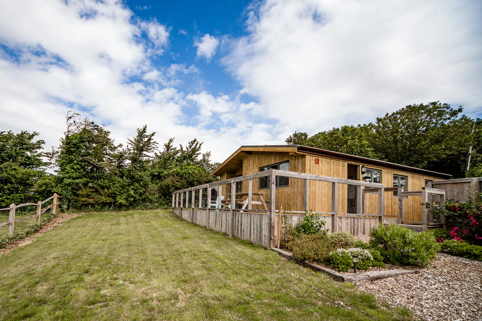 Modern wooden lodge with fenced deck and garden under a blue, partly cloudy sky