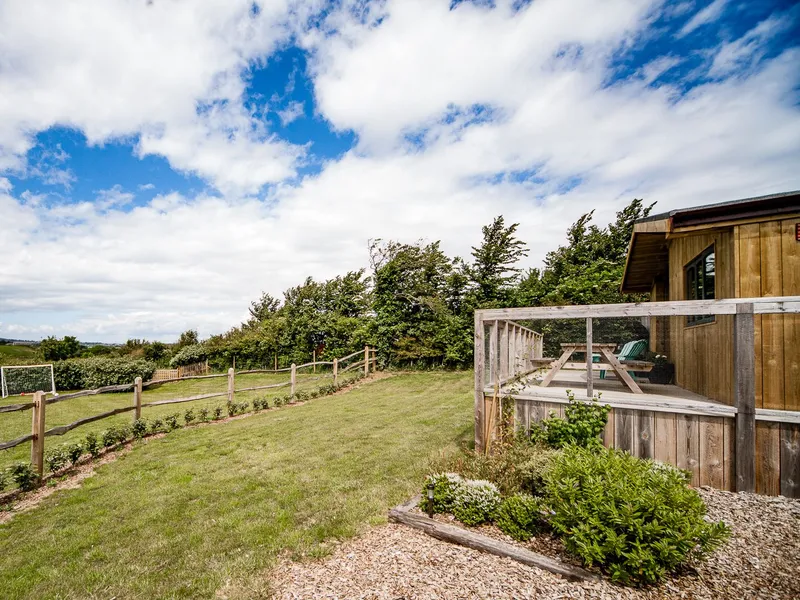 Wooden lodge with enclosed deck overlooking a grassy garden and countryside under a blue, cloud-filled sky