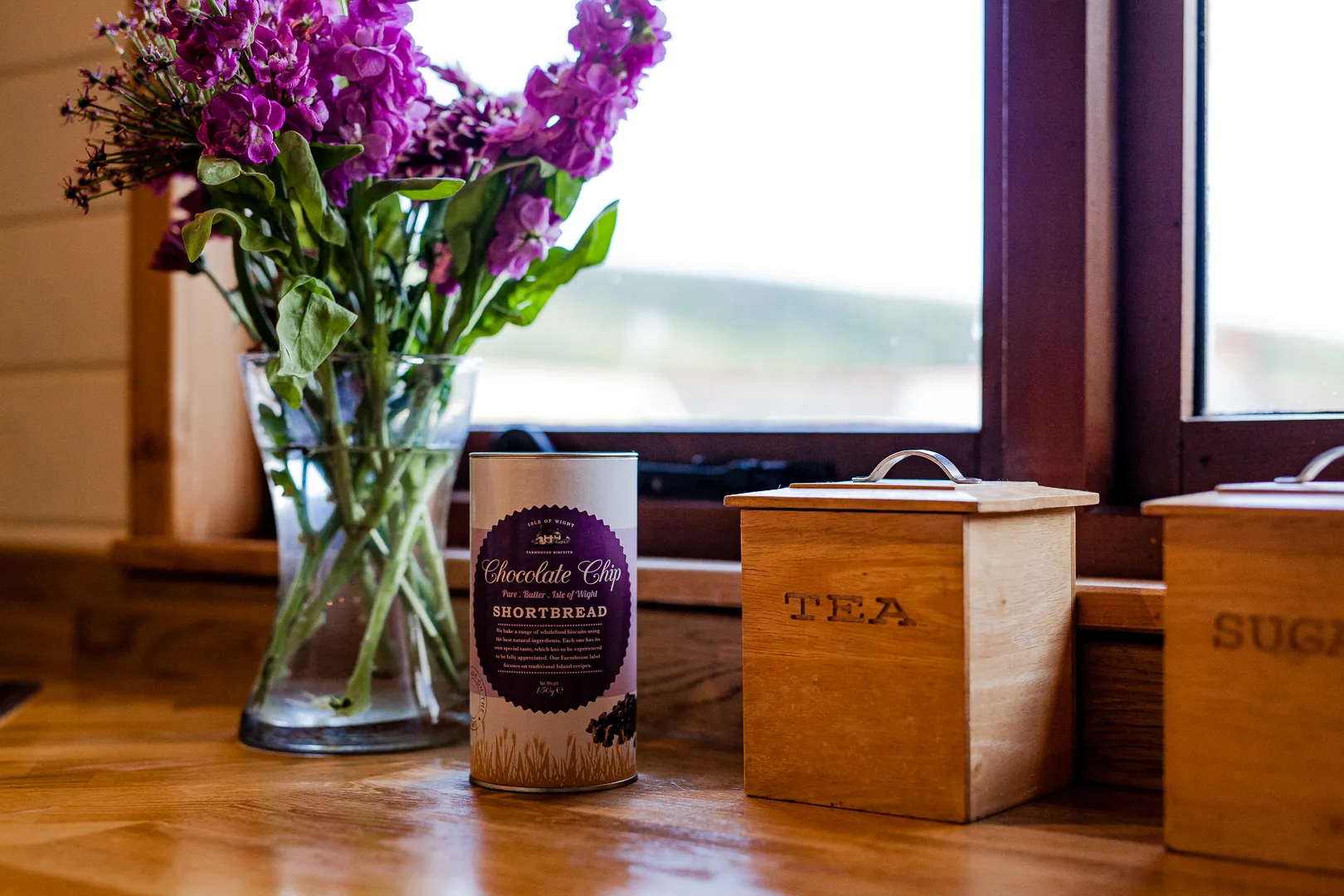 Purple flowers in a glass vase beside a tin of chocolate chip shortbread and wooden tea and sugar canisters on a windowsill