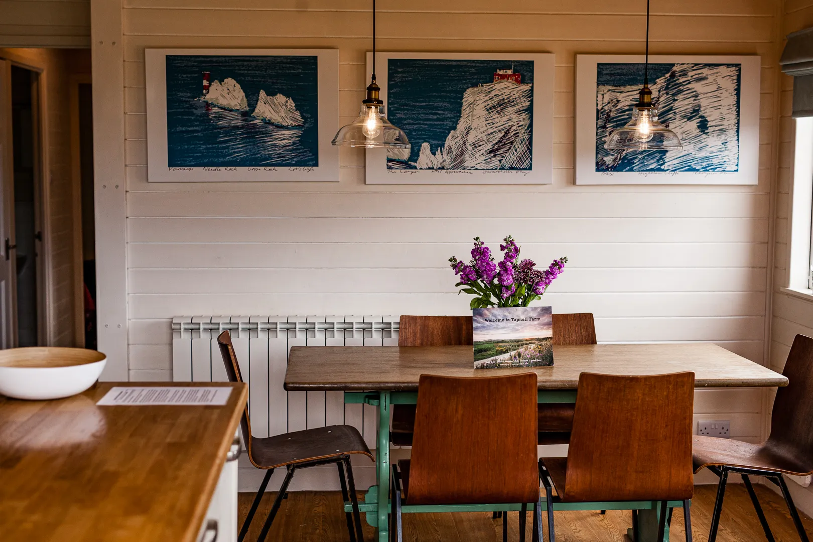 Cosy dining area with wooden table and chairs, pendant lights, framed coastal artwork and a vase of purple flowers