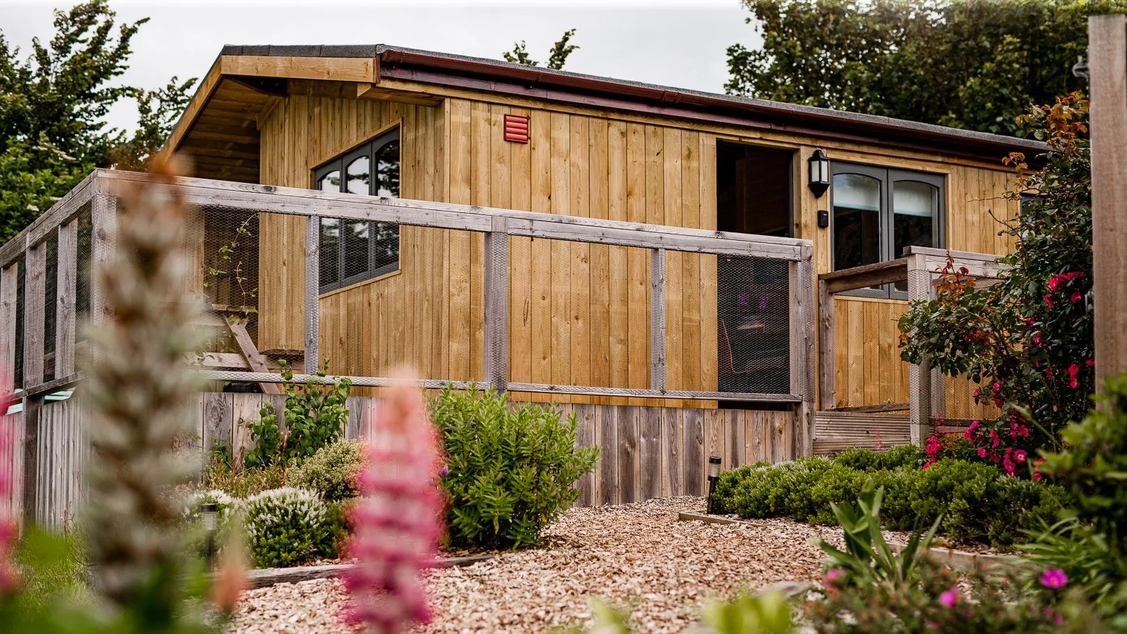 Wooden lodge with a raised deck and mesh balustrade, surrounded by garden plants and flowers