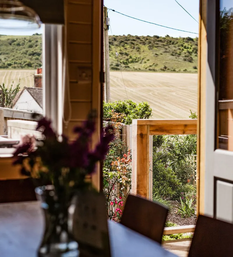 View from a cosy dining area through an open door to a wooden deck and rolling countryside fields
