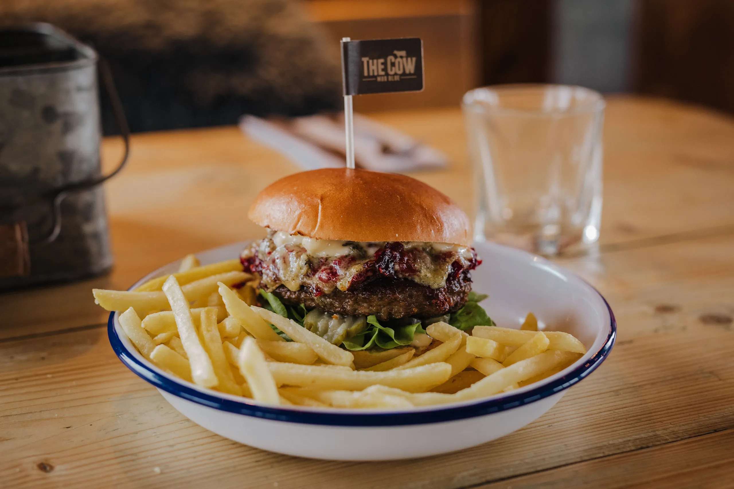 A cheeseburger with lettuce and sauce, served with French fries in a white enamel bowl on a wooden table.