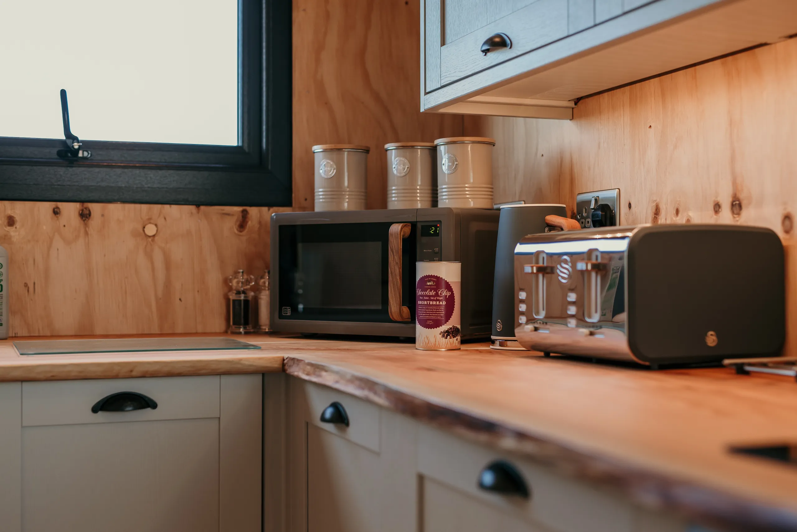 Cosy kitchenette worktop with microwave, toaster, kettle and storage canisters beside a window