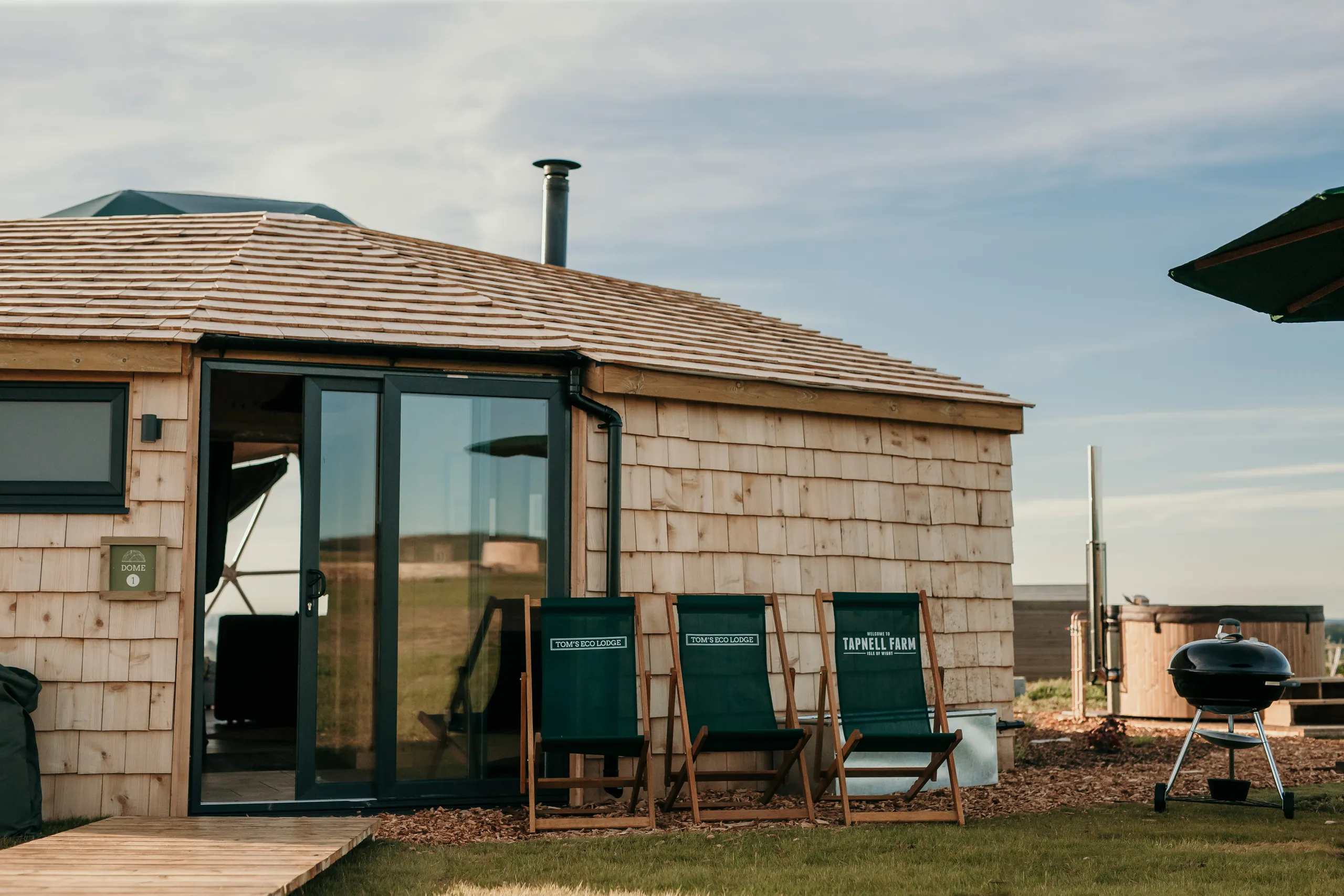 Exterior of a wooden eco lodge with sliding glass doors, three deckchairs and a barbecue beside a hot tub