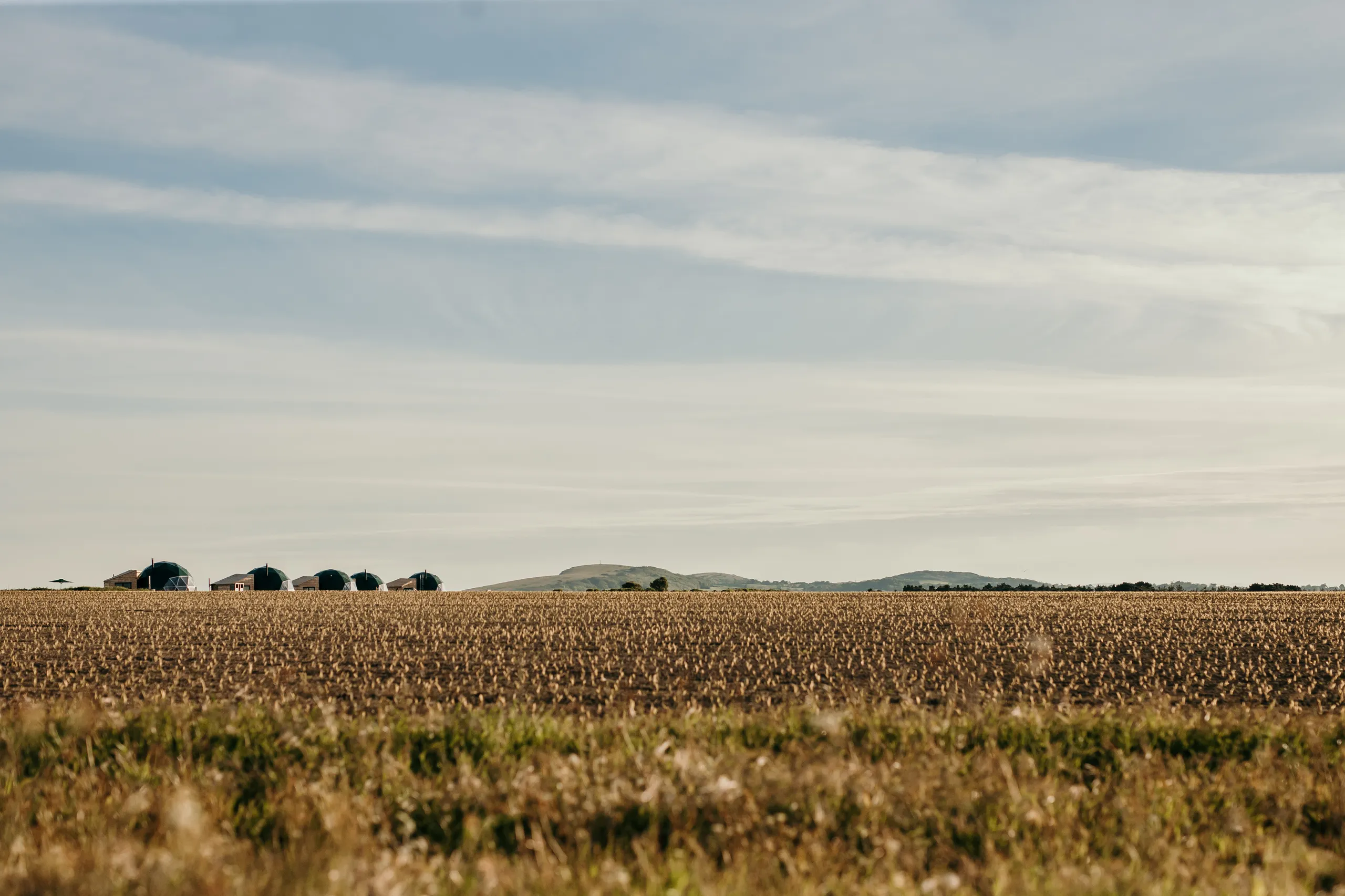 Geodesic dome cabins on the horizon beyond a harvested field under a wide, pale blue sky