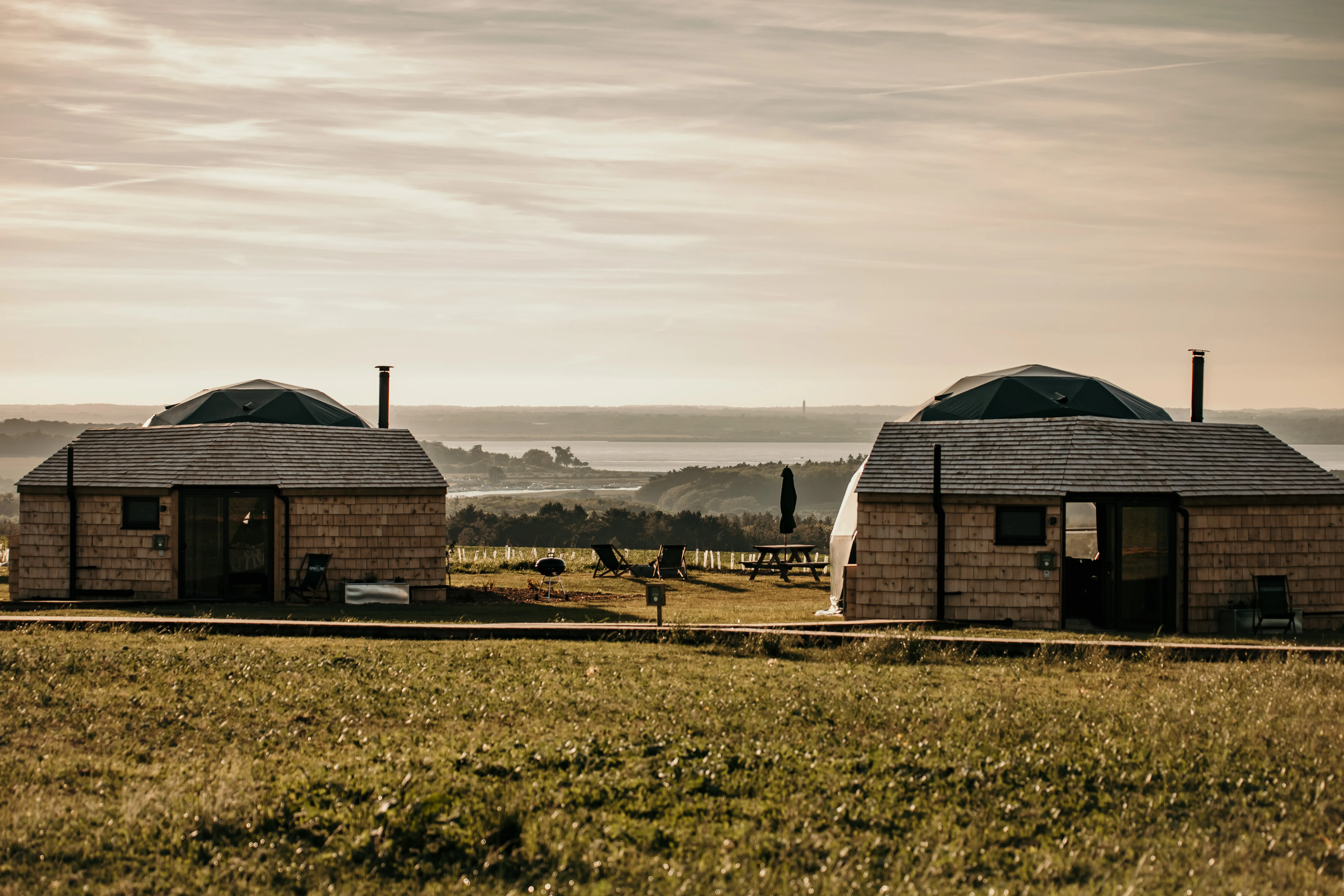 Two wooden glamping cabins with geodesic dome roofs on a grassy hillside overlooking a bay at sunset