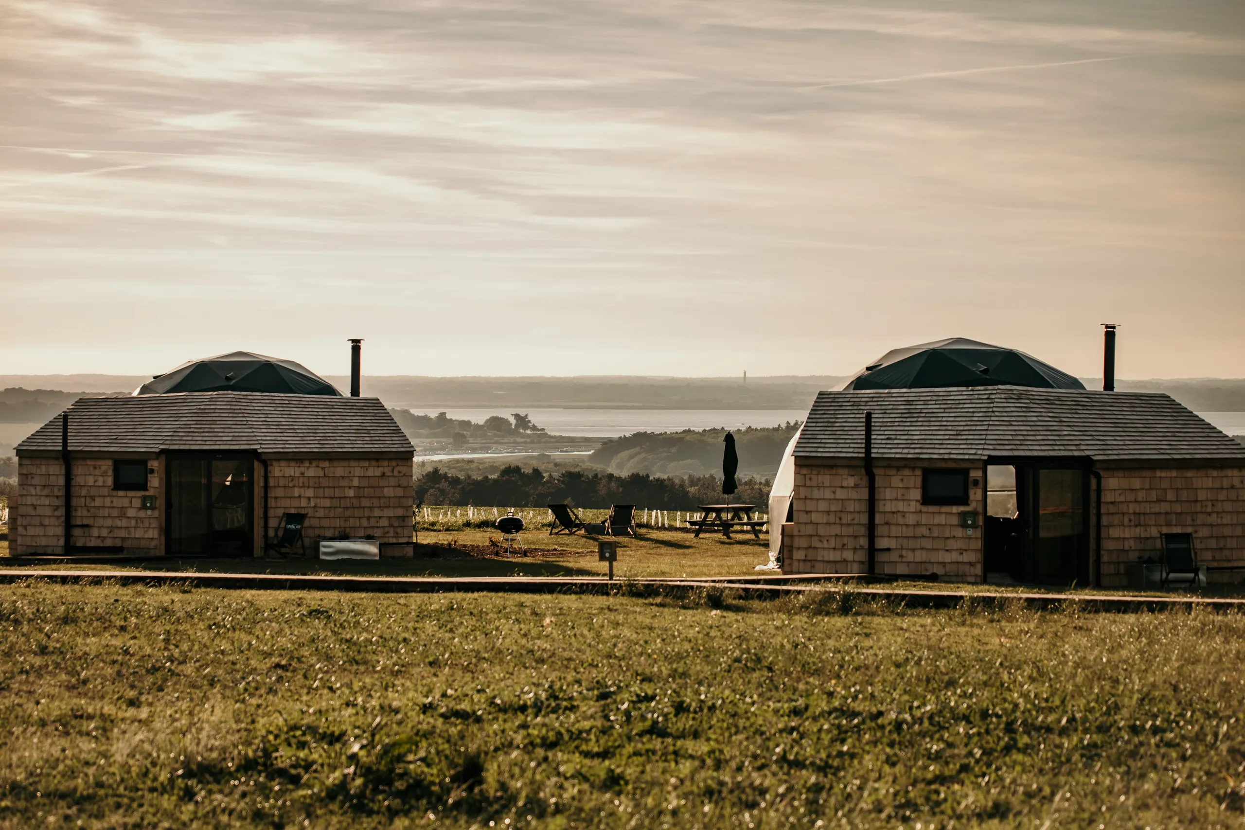 Two wooden glamping cabins with geodesic dome roofs on a grassy hillside overlooking a bay at sunset
