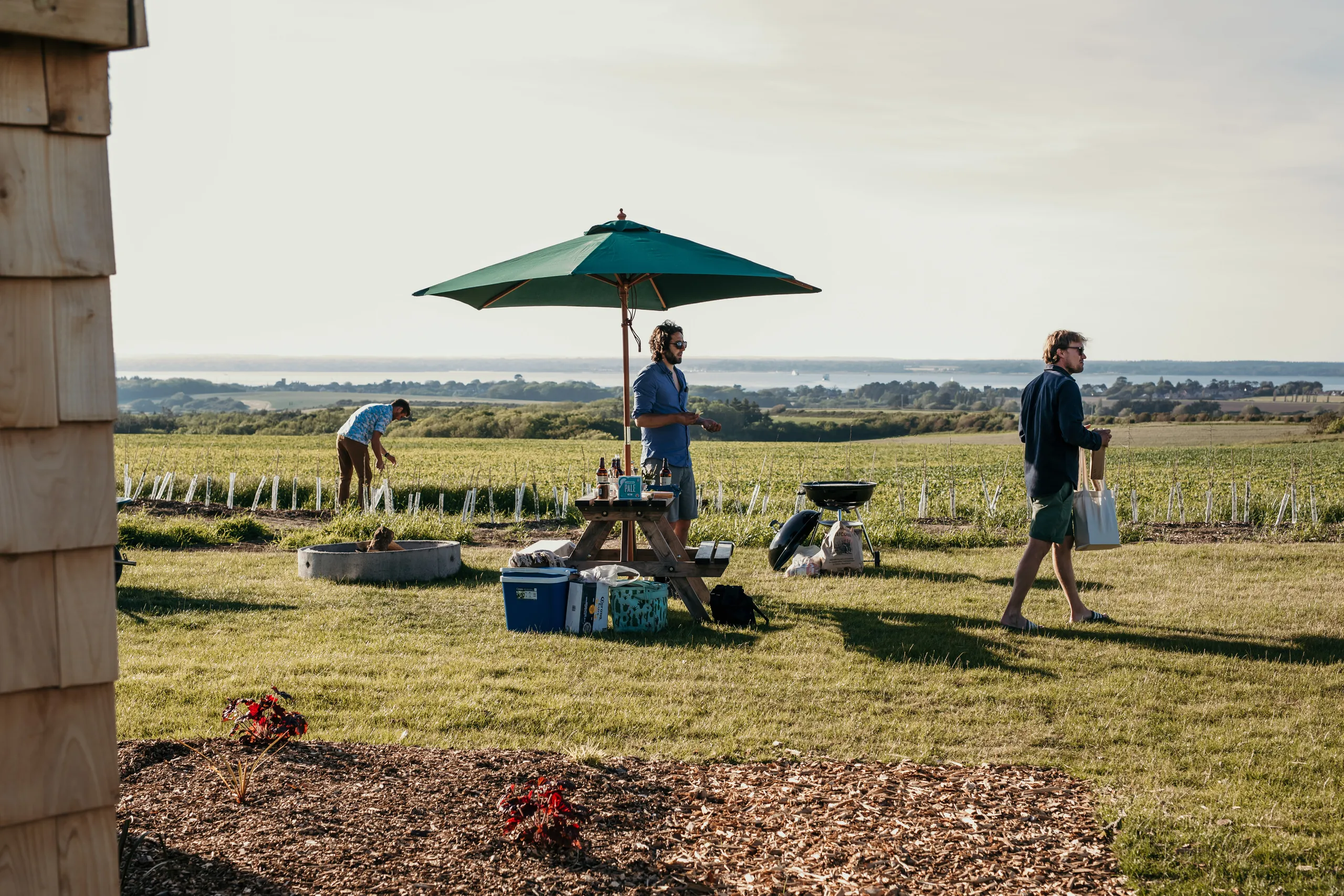 People relaxing by a picnic table under a green parasol in a grassy field with sea views in the distance.