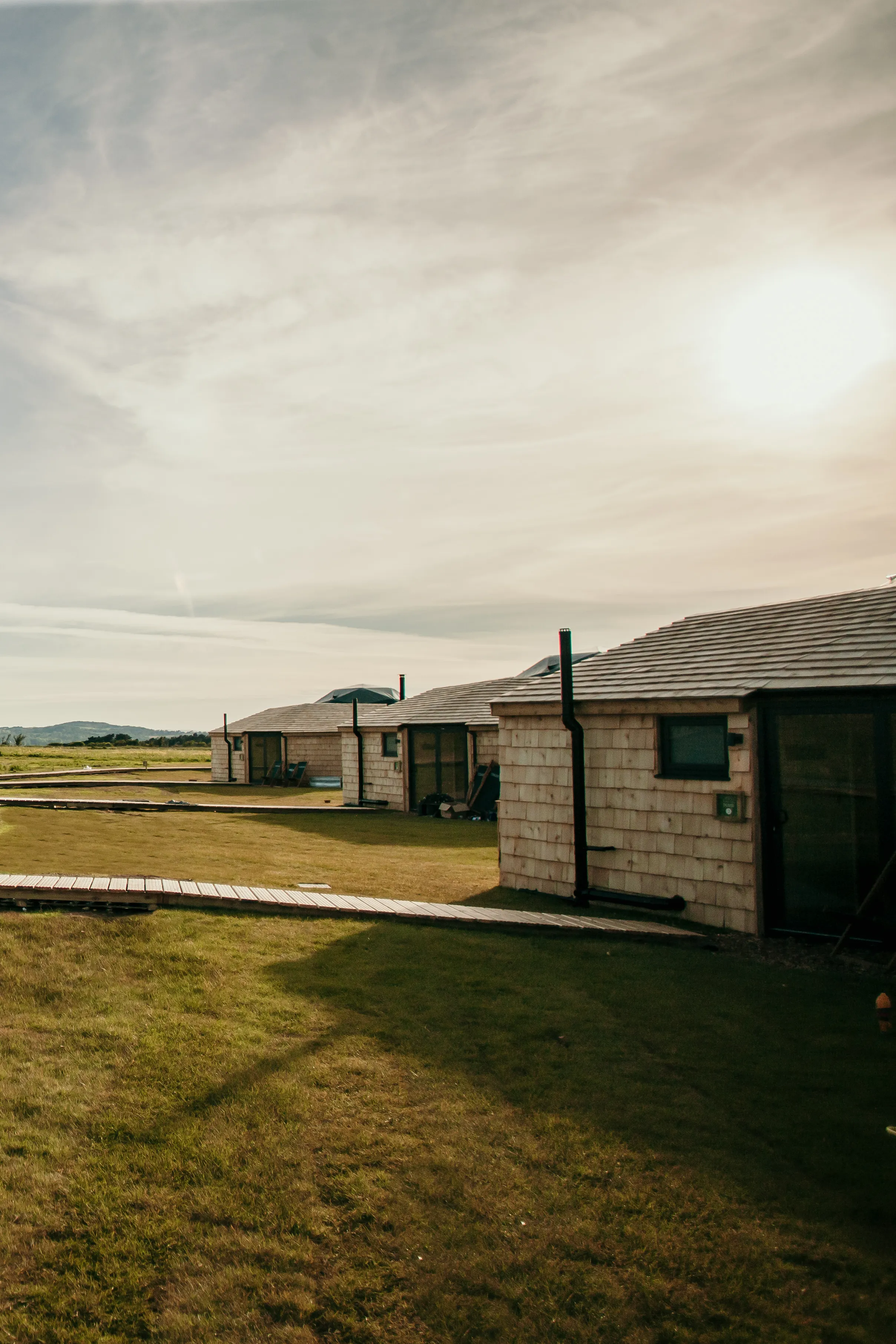 Row of low timber cabins with slate roofs on grassy field in warm evening light