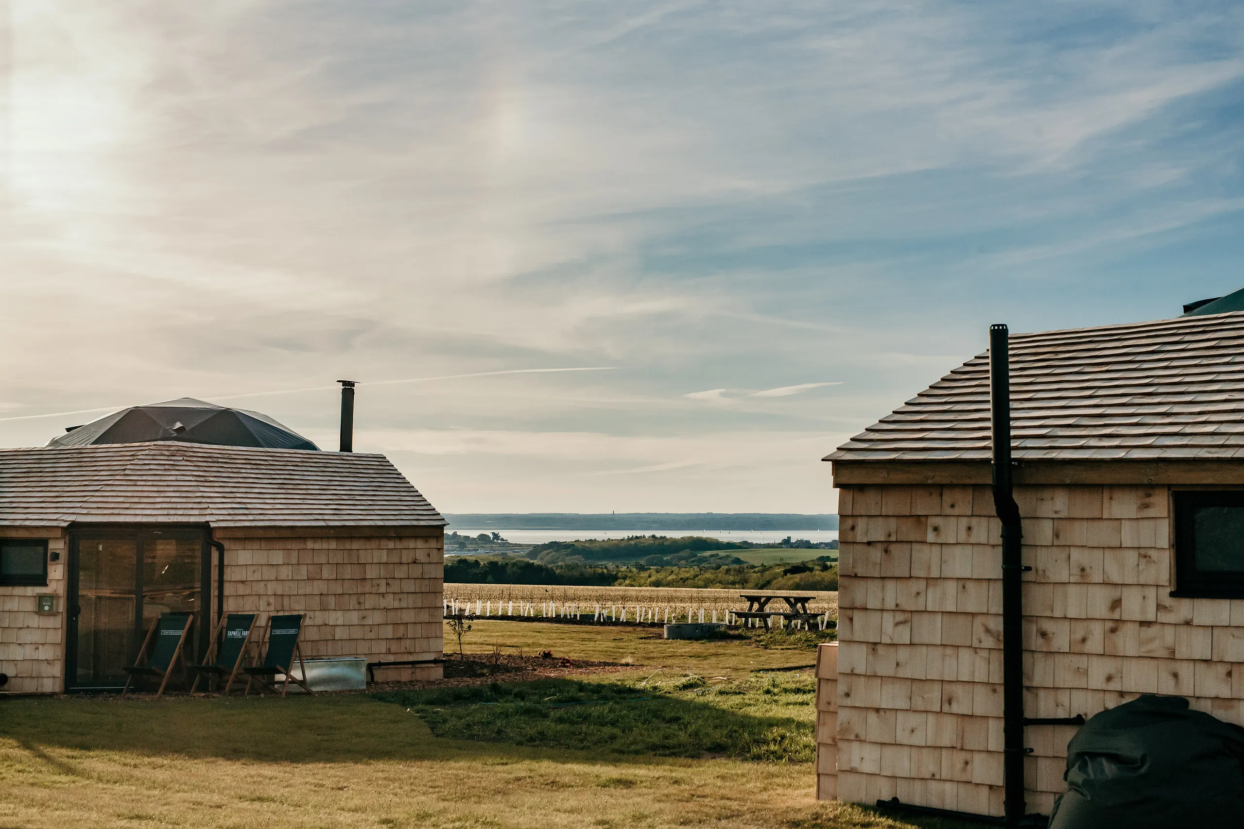 Two wooden cabins with a dome roof and chimney overlooking fields and the sea at sunset