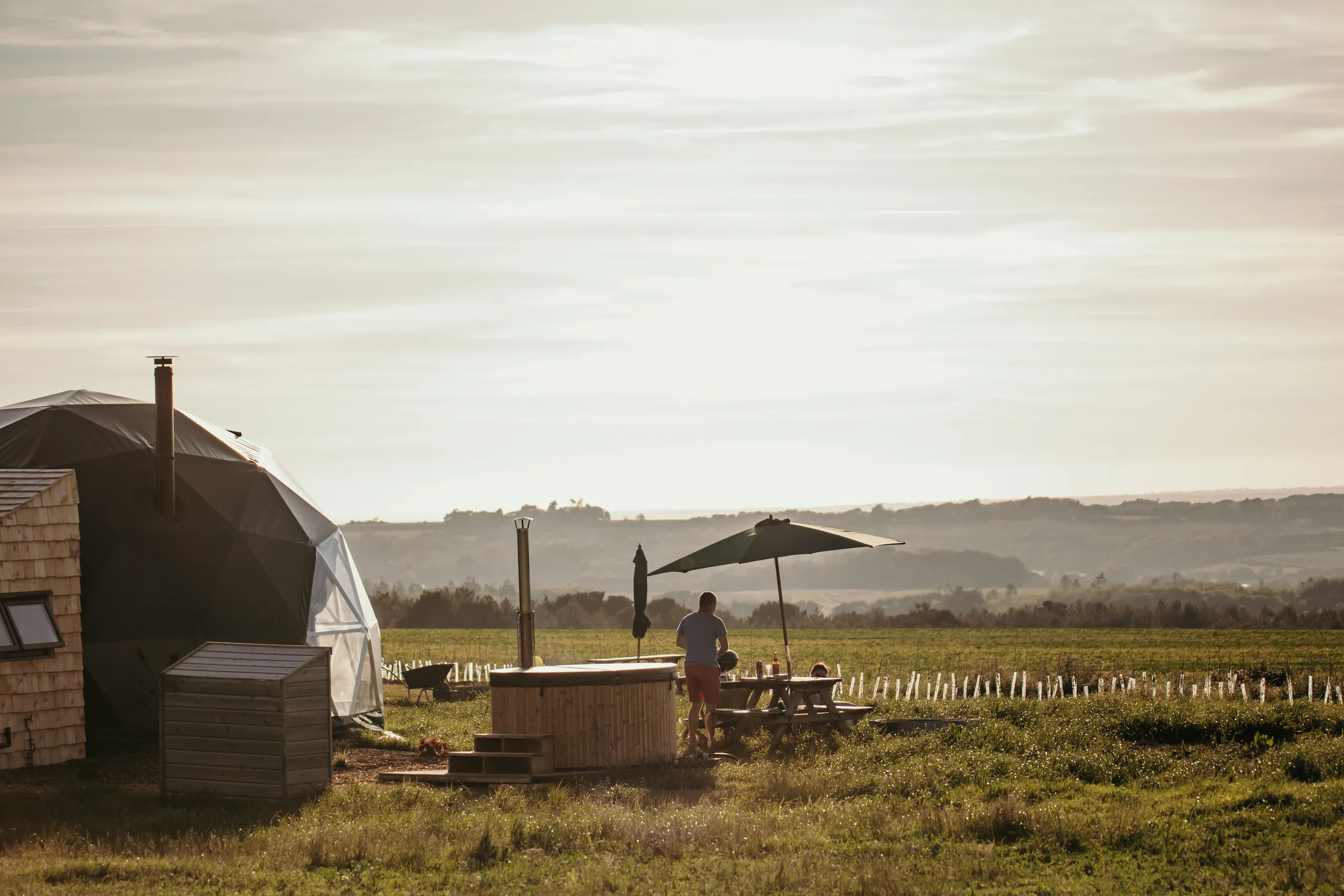 Geodesic dome glamping pod beside a wooden hot tub in a grassy field at sunset