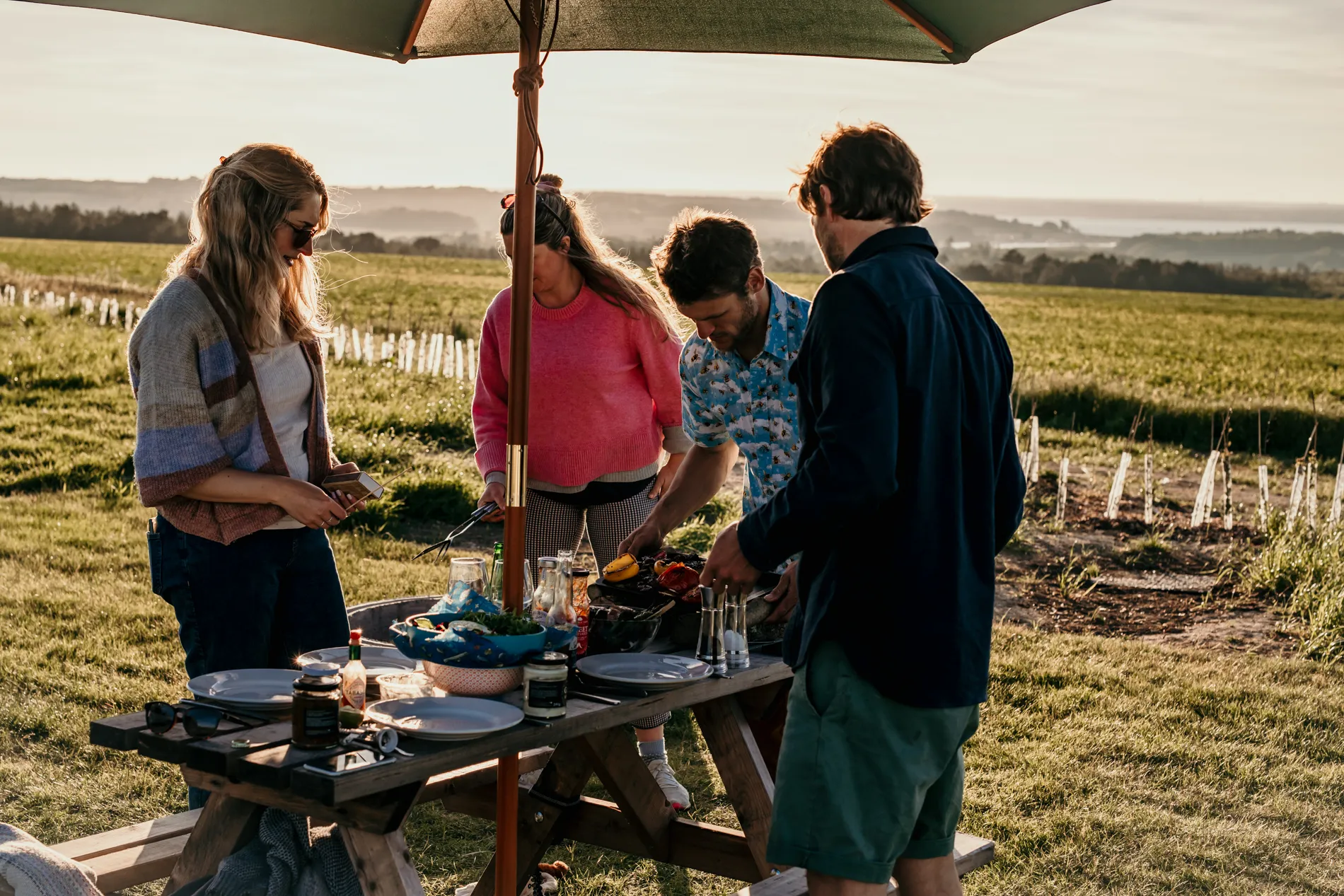 Group of friends preparing a barbecue meal at a picnic table under a parasol in a countryside field at sunset