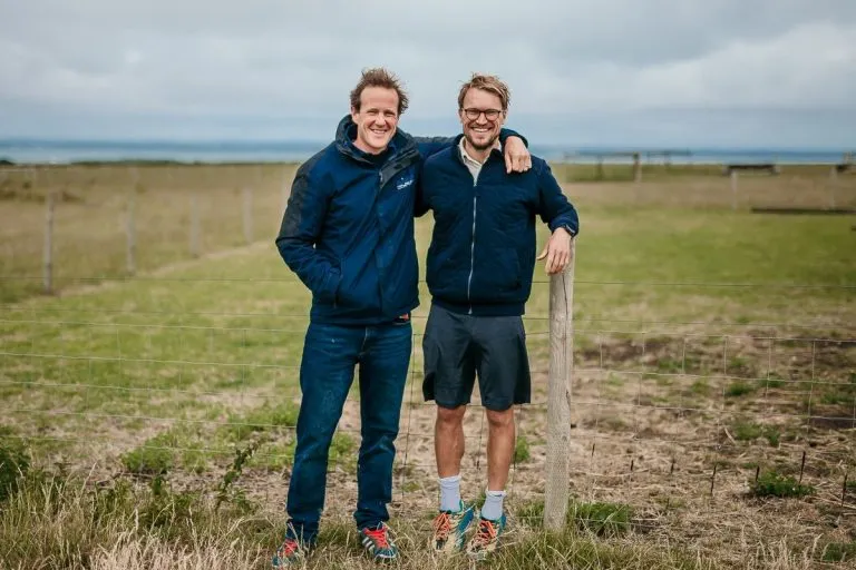 Two men standing together in a field, one with his arm around the other's shoulder, both smiling at the camera