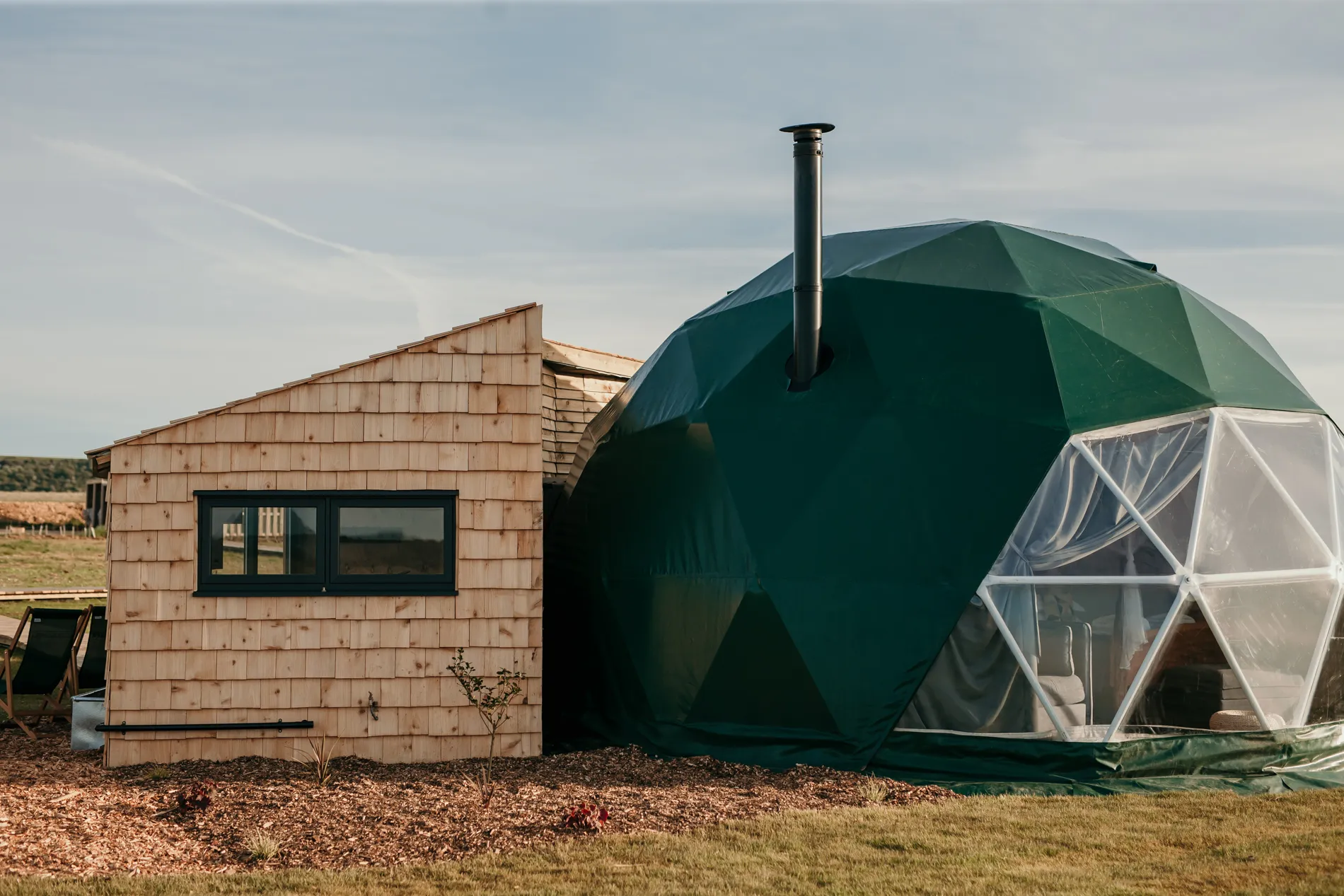 Green geodesic glamping dome with large window and chimney beside a wooden cabin on open countryside