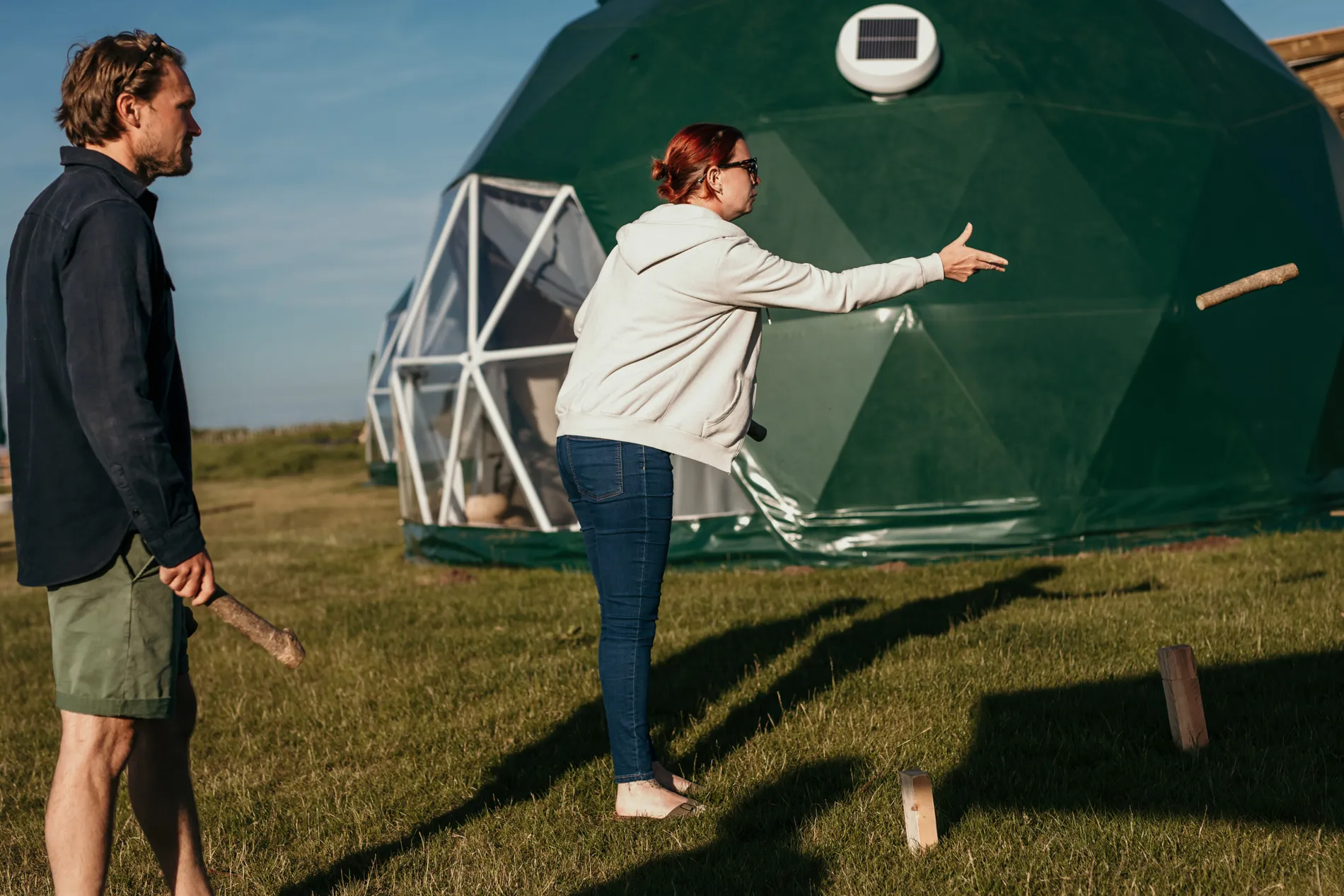 Woman tossing a wooden baton during an outdoor game beside green geodesic dome tents, with a man watching on the grass.