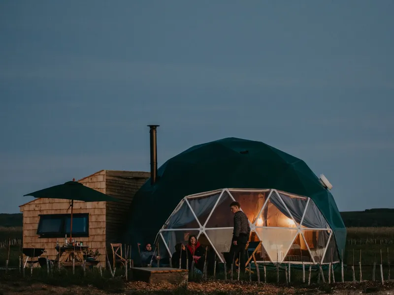 Family relaxing outside a geodesic dome with warm lights at dusk in a rural field