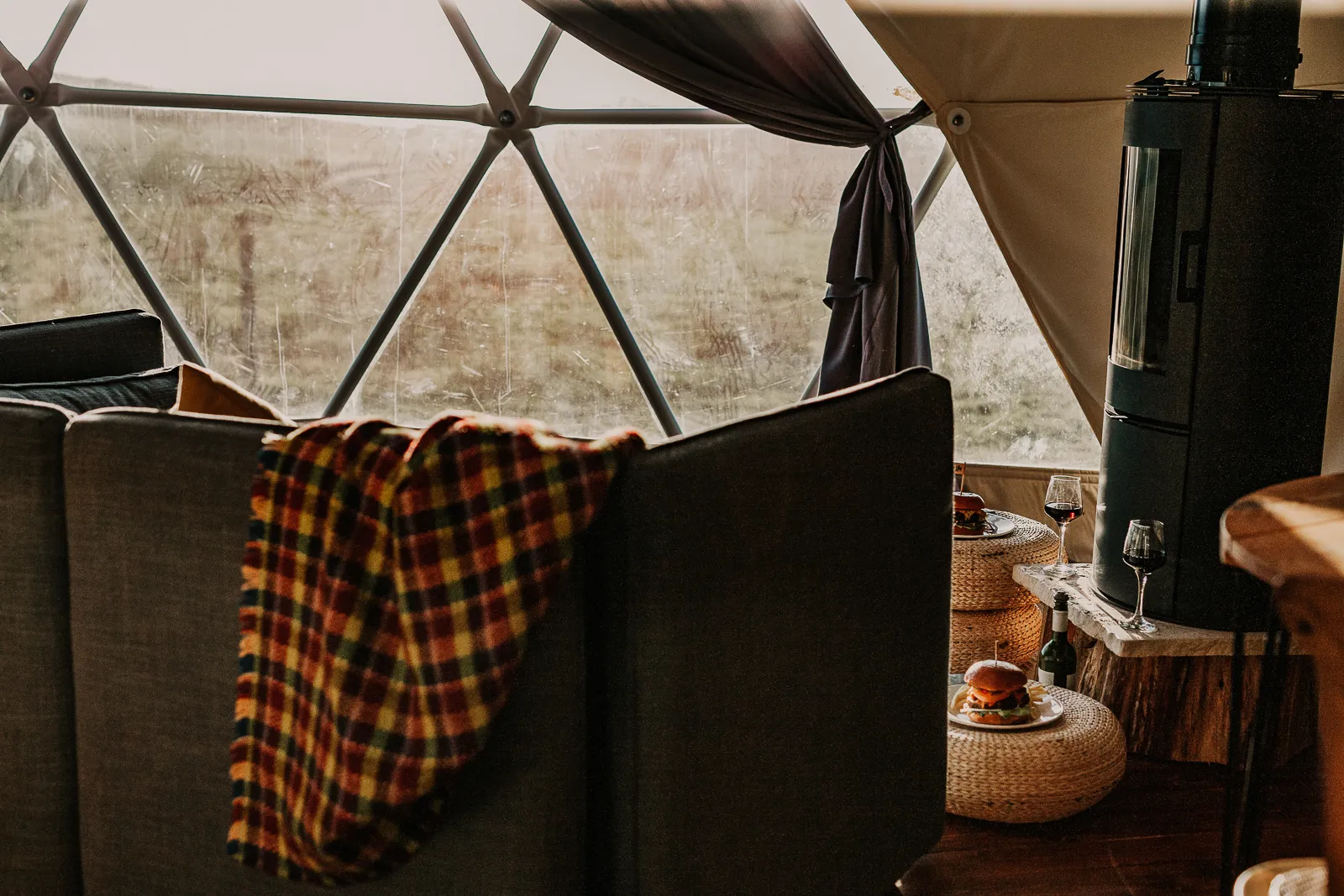 Cosy geodesic dome interior with a sofa, tartan blanket and wood-burning stove beside small tables with wine and food