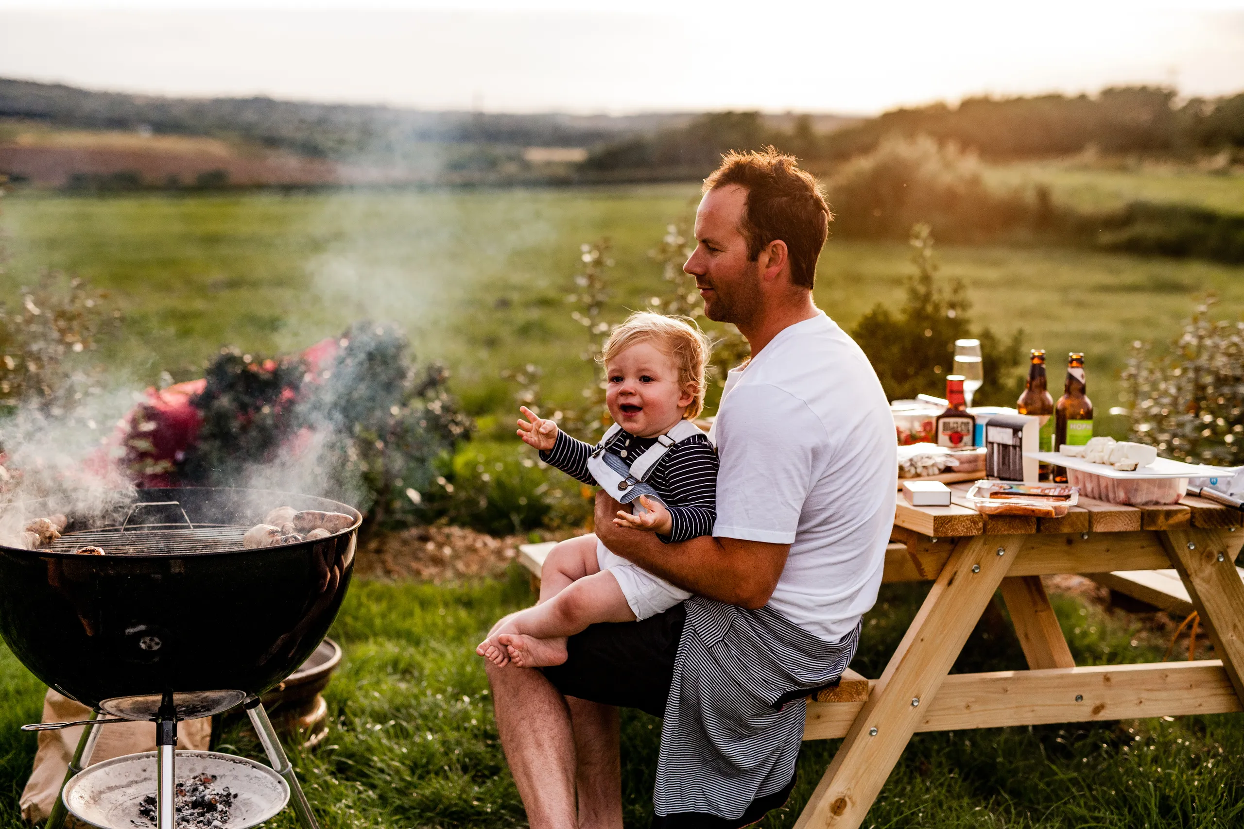 Adult holding a toddler beside a smoky barbecue grill at an outdoor picnic table in the countryside at sunset