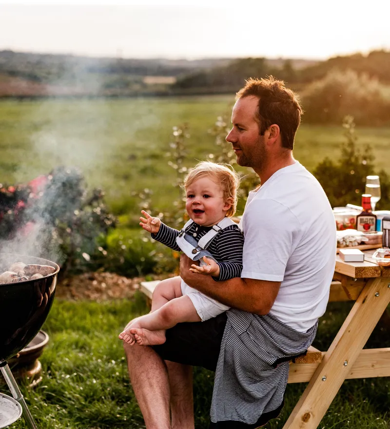 Adult holding a toddler beside a smoky barbecue grill at an outdoor picnic table in the countryside at sunset