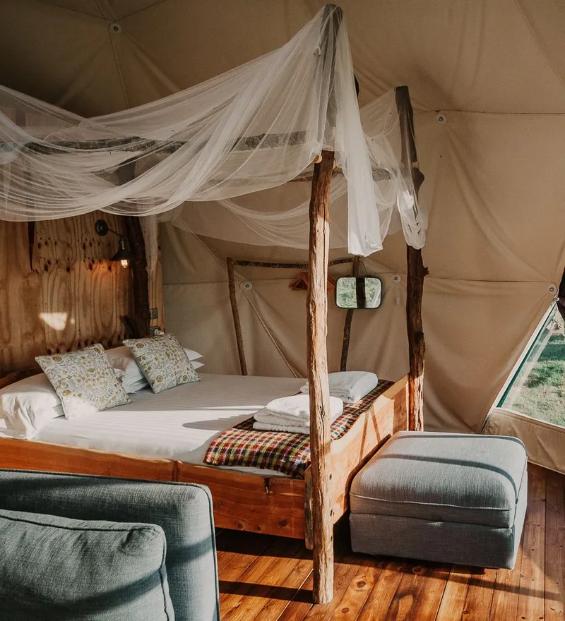 Cosy glamping dome interior with wooden four-poster bed, draped netting and large window overlooking countryside