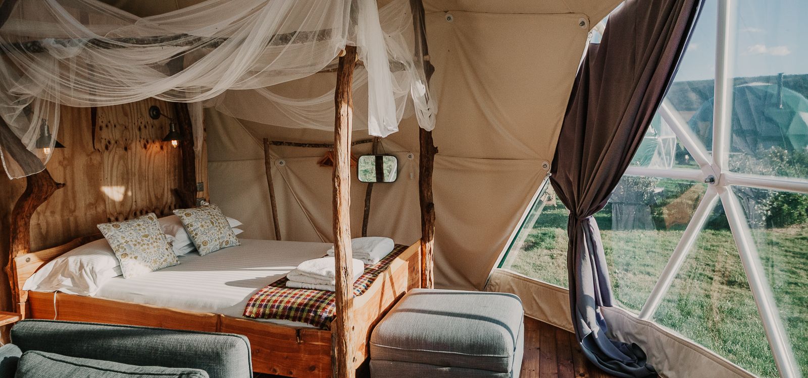 Cosy glamping dome interior with wooden four-poster bed, draped netting and large window overlooking countryside
