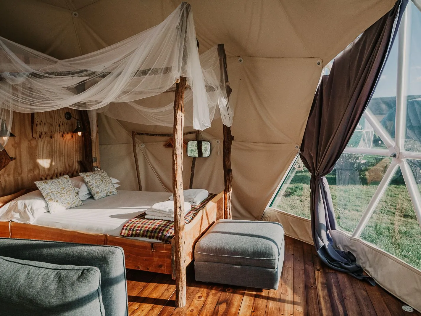 Cosy glamping dome interior with wooden four-poster bed, draped netting and large window overlooking countryside