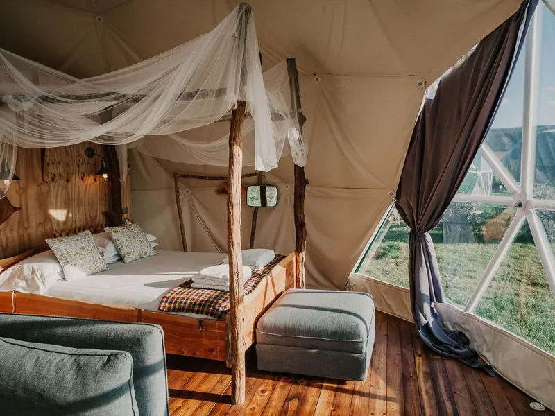 Cosy glamping dome interior with wooden four-poster bed, draped netting and large window overlooking countryside