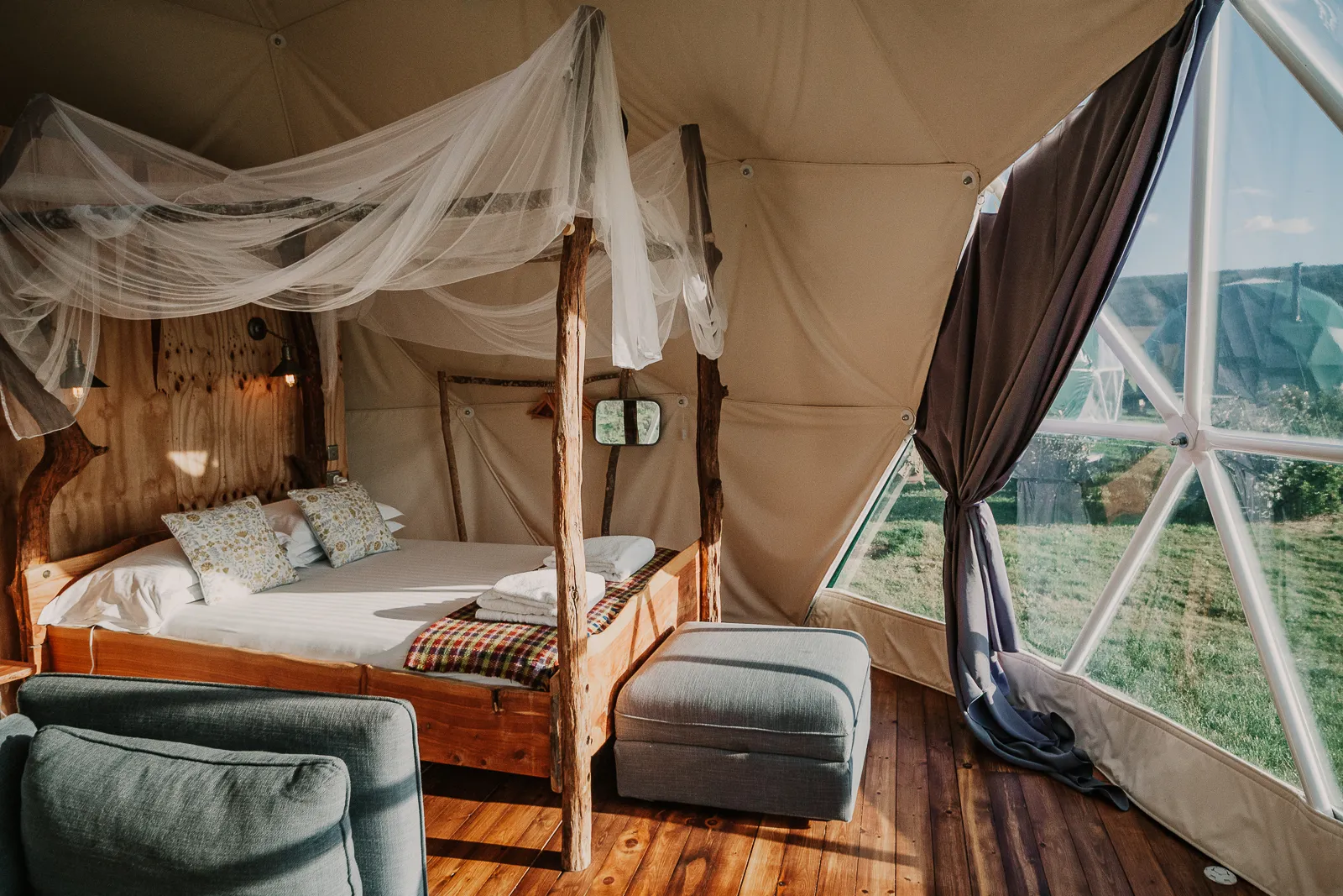 Cosy glamping dome interior with wooden four-poster bed, draped netting and large window overlooking countryside