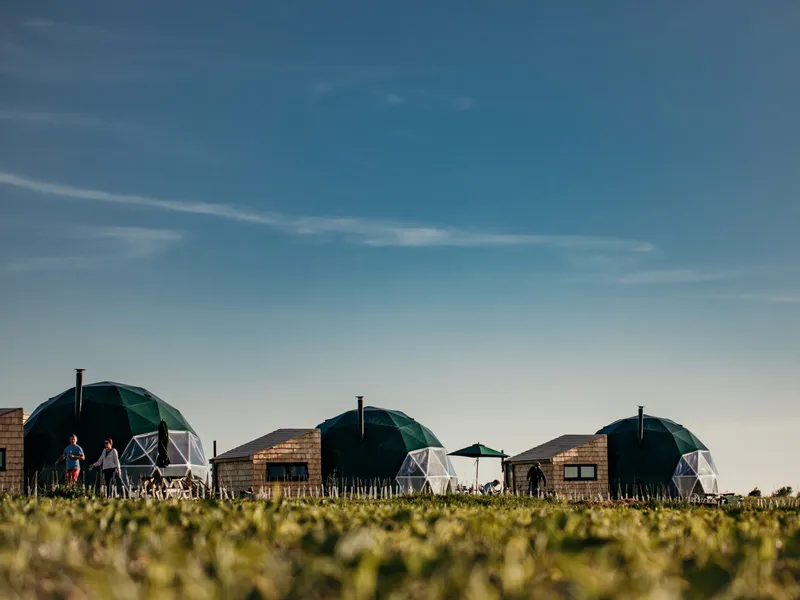 Geodesic dome cabins and small wooden huts on a grassy field under a wide blue sky