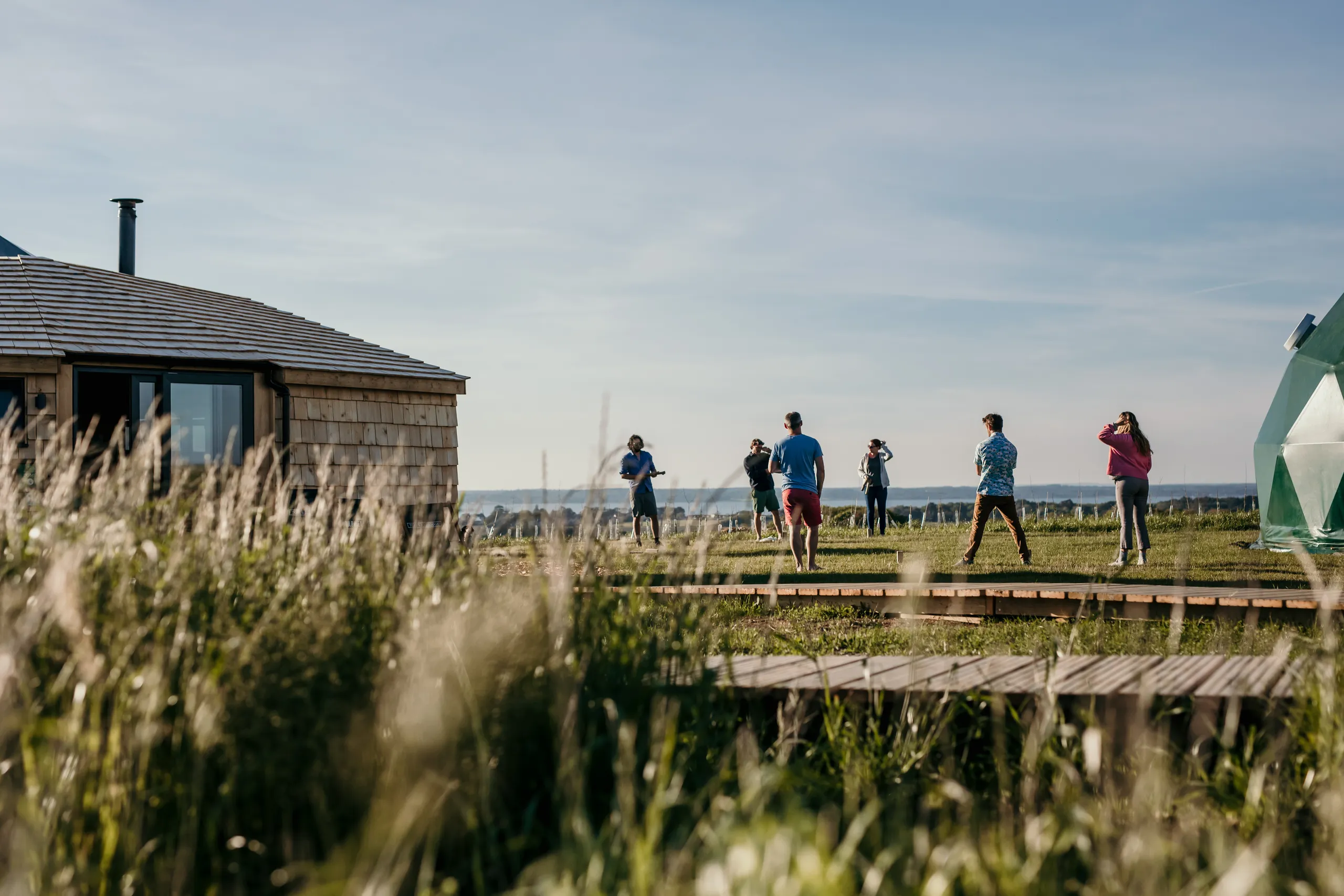 People standing on a grassy field beside a wooden cabin and a green geodesic dome under a blue sky