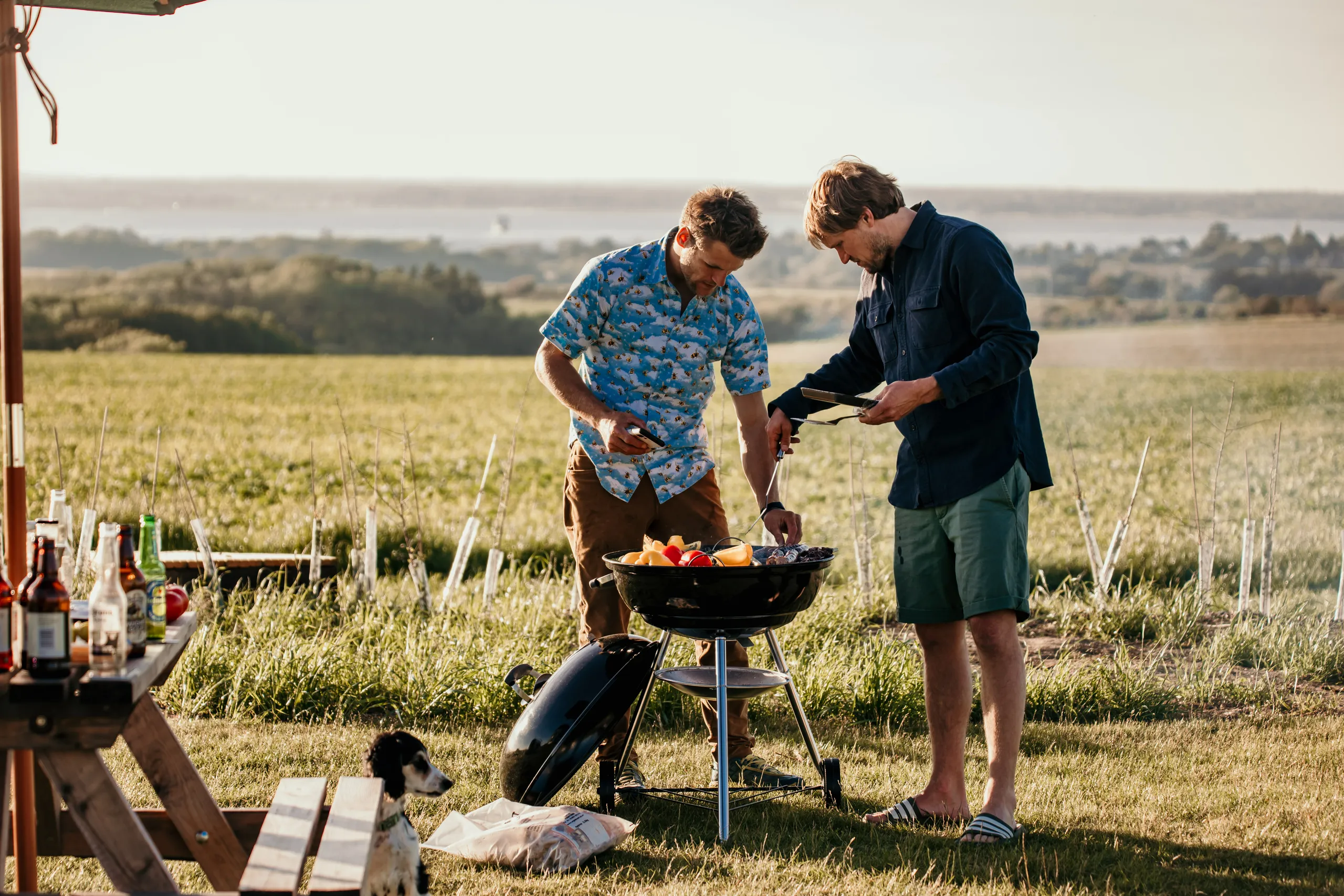 Two men grilling food on a barbecue in a grassy field with countryside views