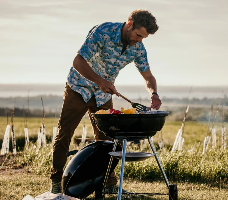 Man grilling vegetables on a barbecue in a grassy field at sunset