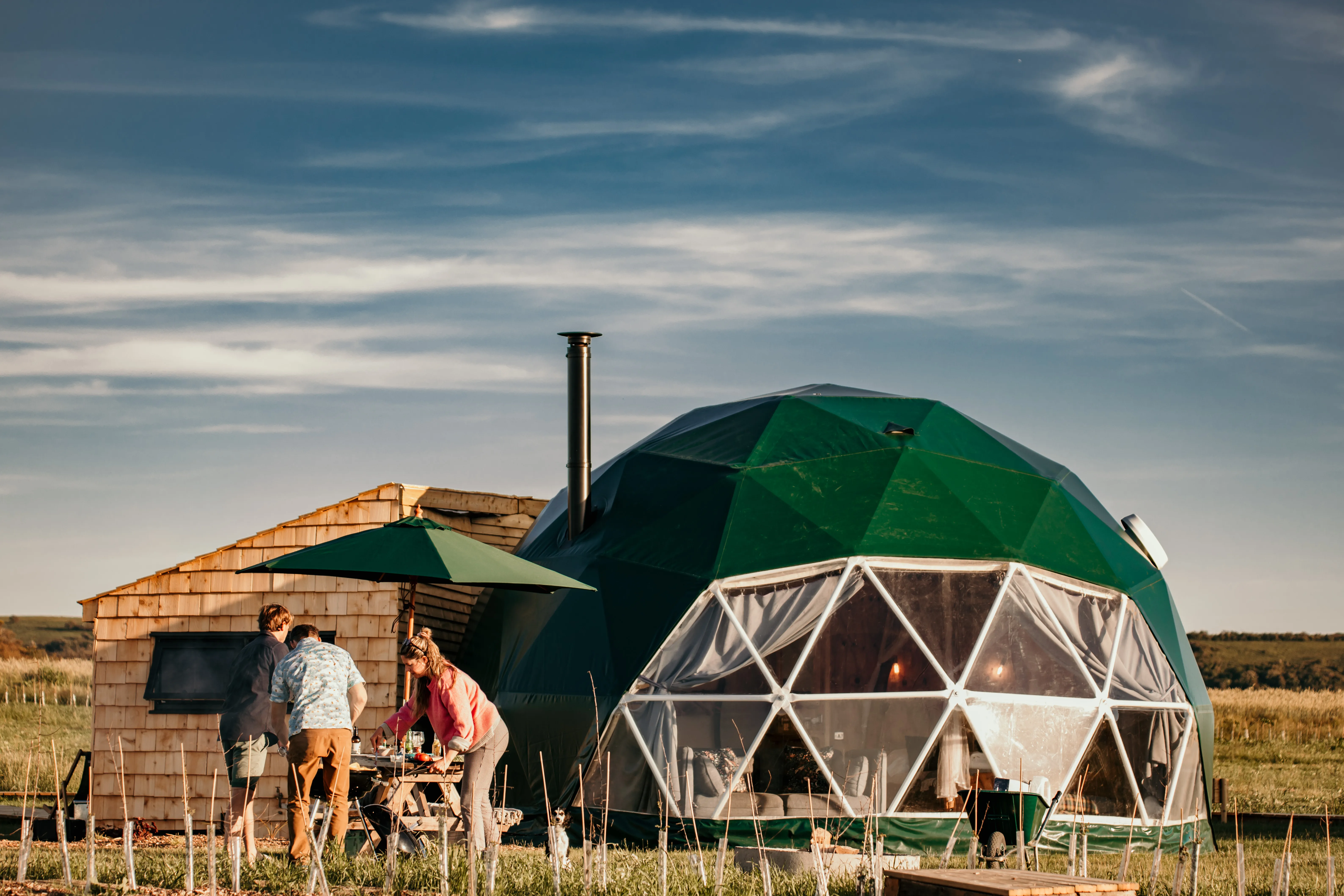 People preparing a barbecue outside a green geodesic dome with a chimney in a rural field