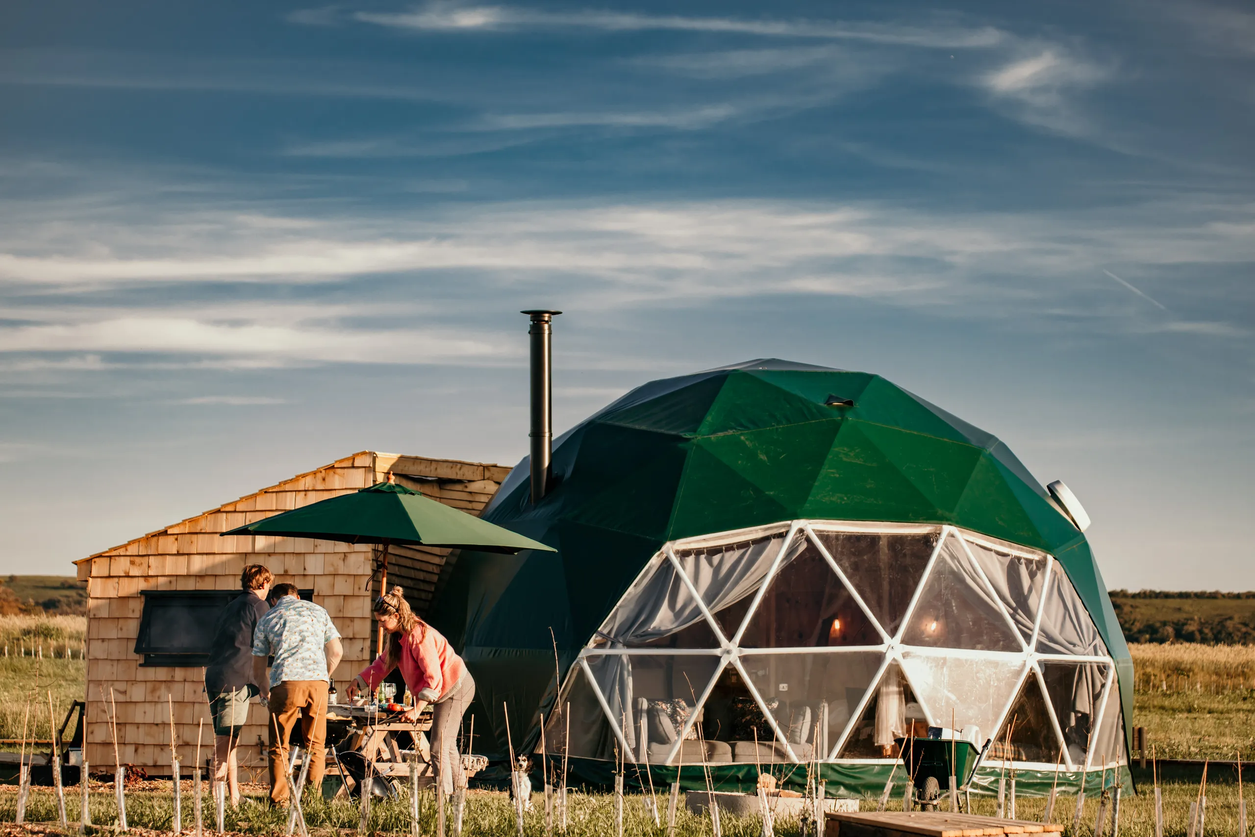 People preparing a barbecue outside a green geodesic dome with a chimney in a rural field